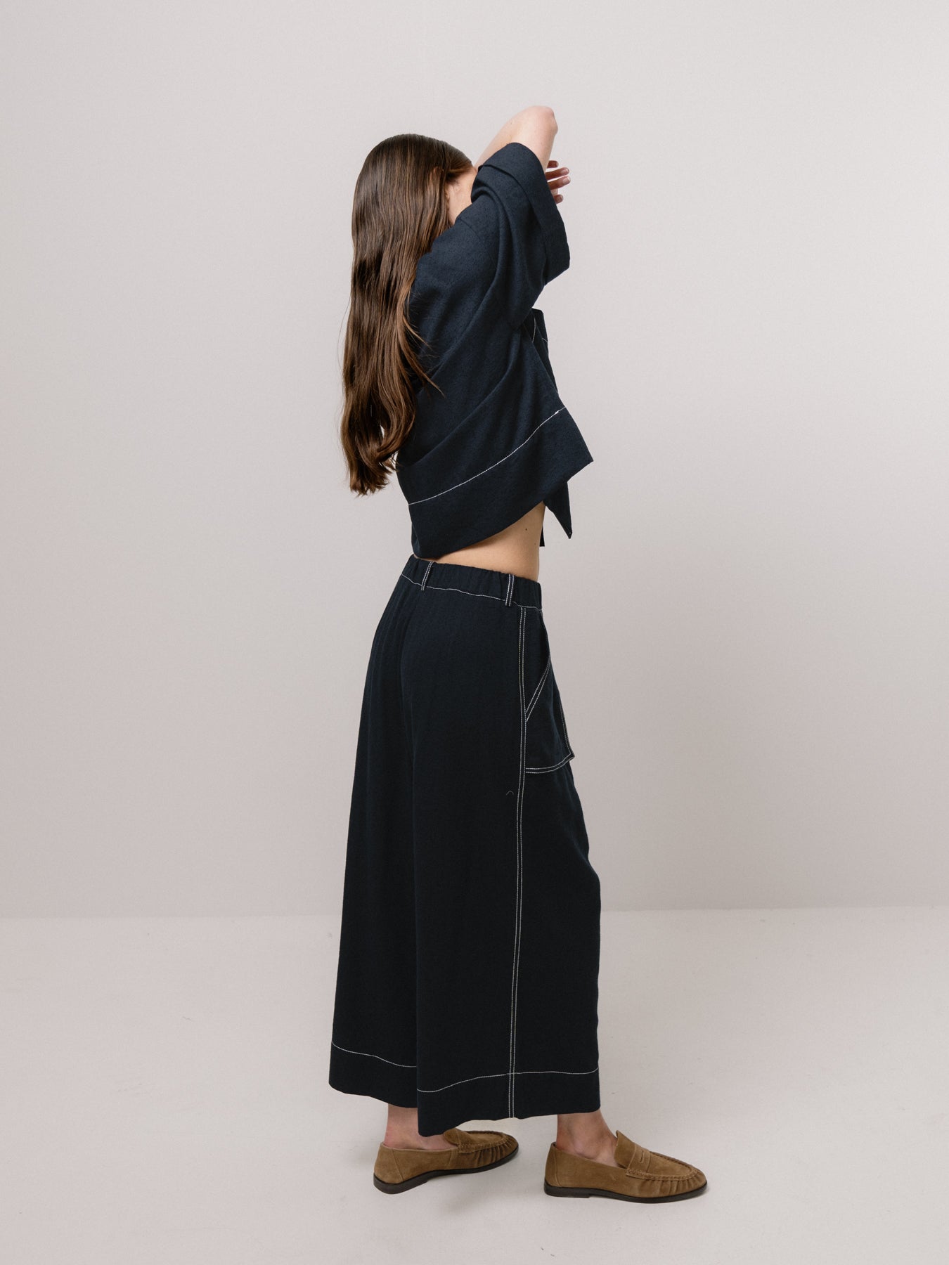 A woman in a dark outfit exudes effortless style and elegance, standing sideways with arms raised against a plain background. She wears Emin and Paul's Wide Leg Trousers - Black, paired with a top and brown shoes.