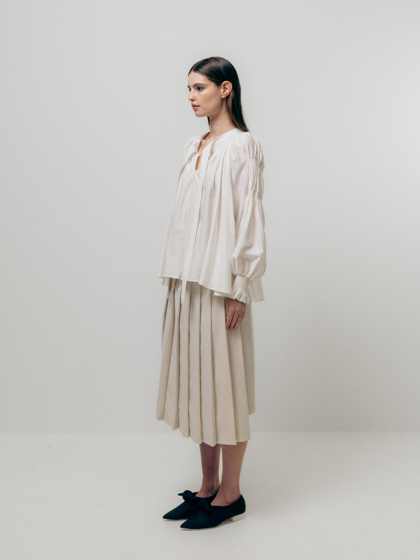 A woman in an Emin and Paul Shirring Sleeve Blouse - White, standing in profile against a plain background, pairs this versatile wardrobe staple with a pleated skirt and black shoes, effortlessly blending style and comfort.