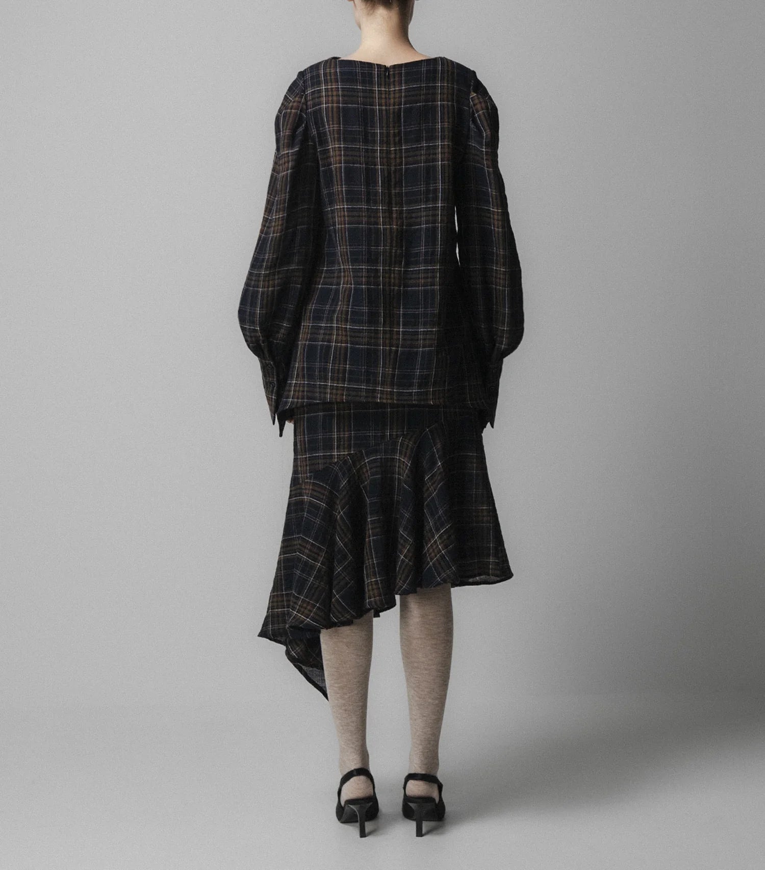 A person stands facing away against a plain grey background, wearing the Emin and Paul Volume Sleeve Blouse in Navy Check with oversized sleeves, an asymmetrical skirt, light tights, and black heeled sandals.