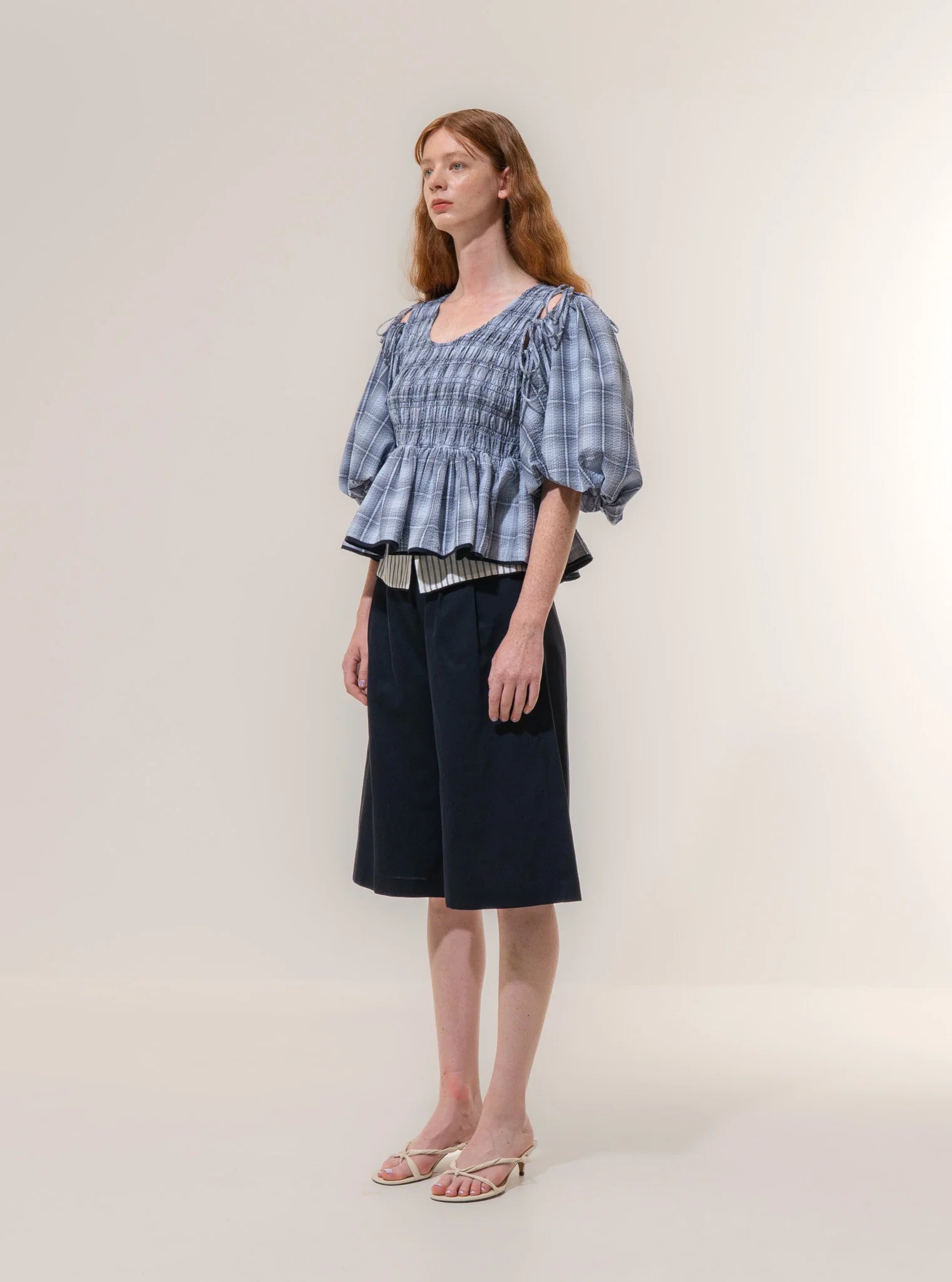A woman stands against a plain background wearing the Emin and Paul Puff Sleeve Tie Blouse - Blue Check, styled with black knee-length shorts and white sandals. The 100% cotton blouse adds comfort to her effortlessly chic look.