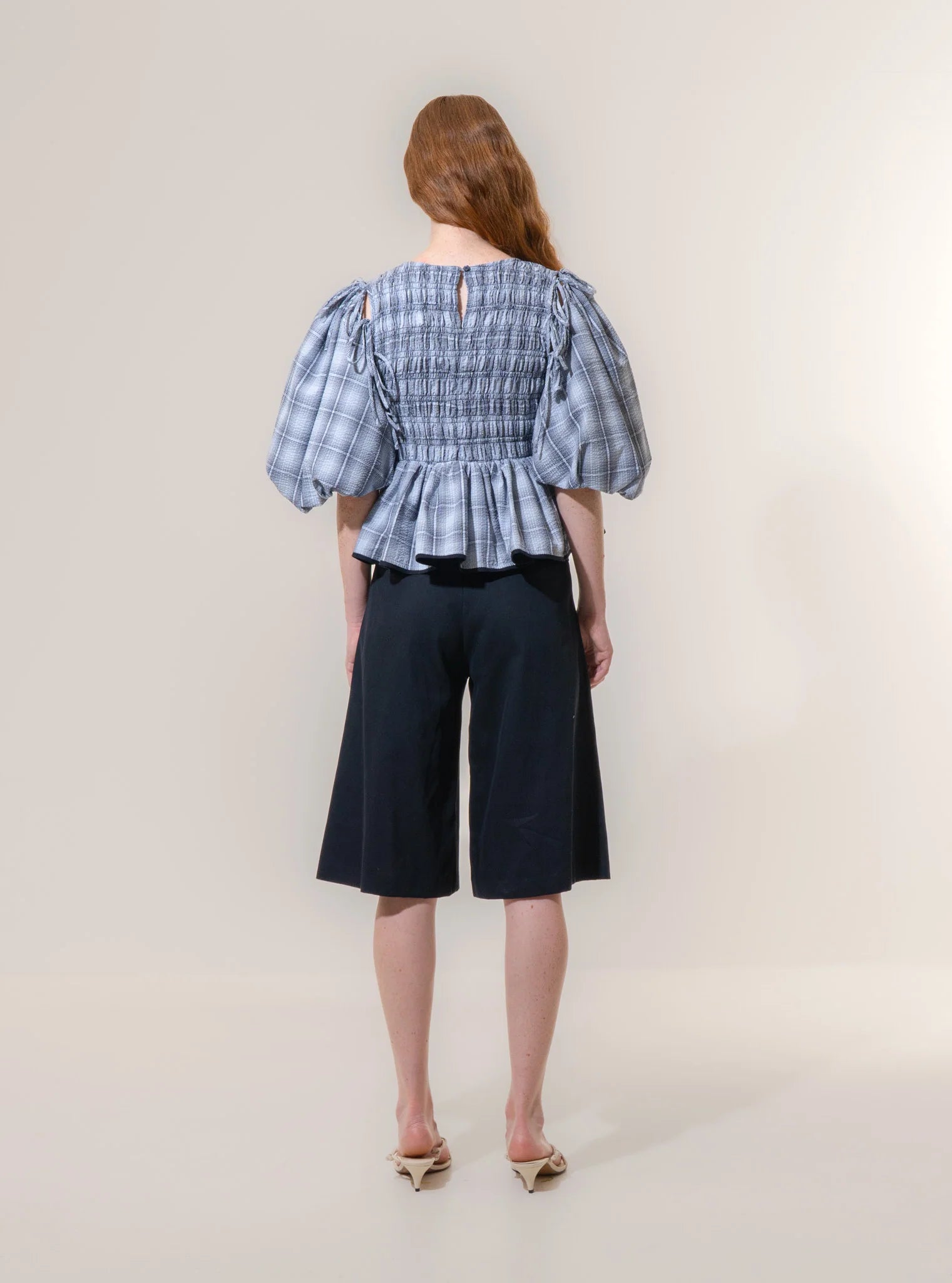 A person faces away, wearing the Emin and Paul Puff Sleeve Tie Blouse - Blue Check with black knee-length shorts and beige pointed-toe shoes against a plain background.