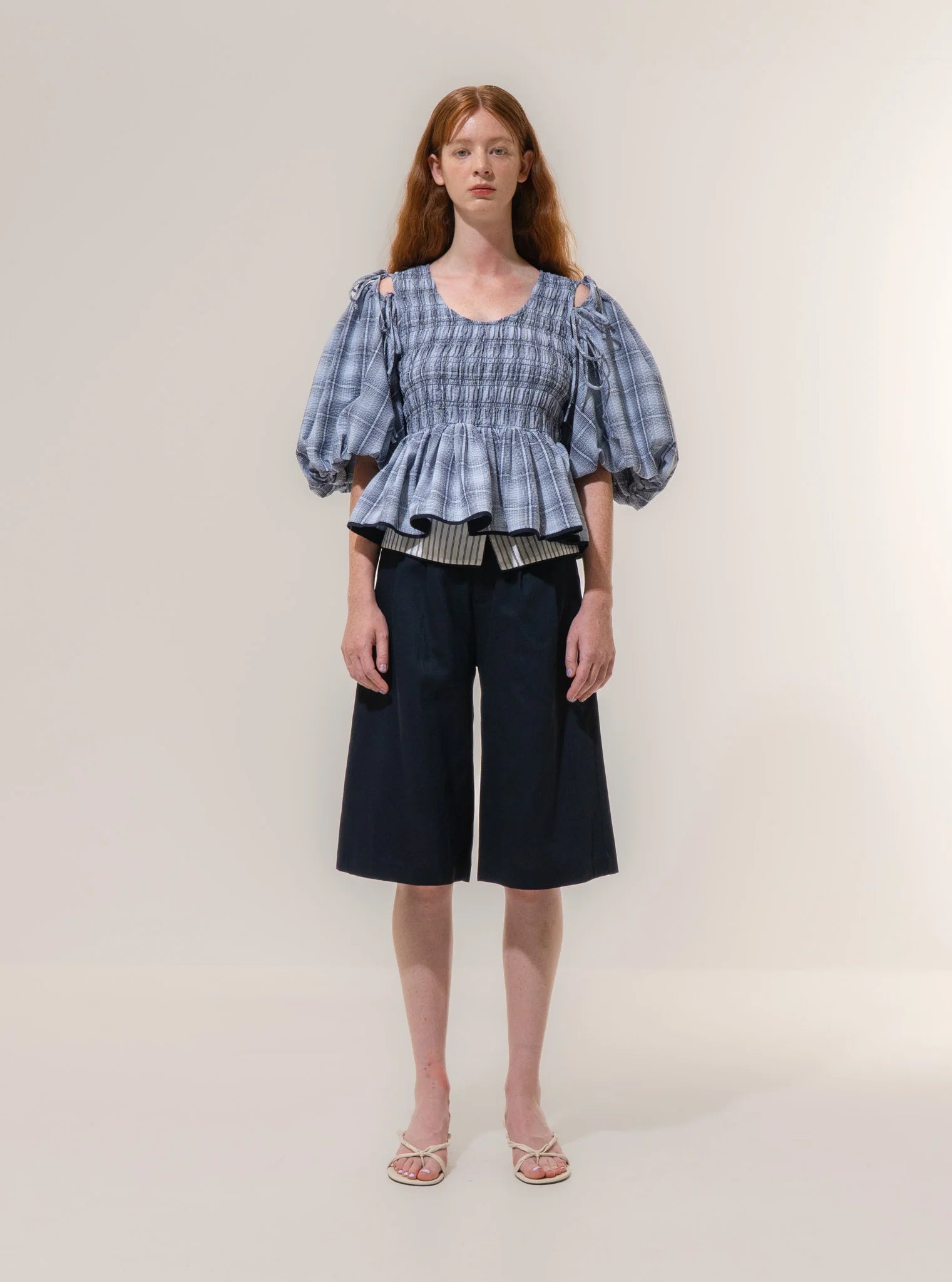 A person stands facing forward wearing the Emin and Paul Puff Sleeve Tie Blouse - Blue Check, matched with knee-length navy shorts and white open-toe shoes against a plain background.