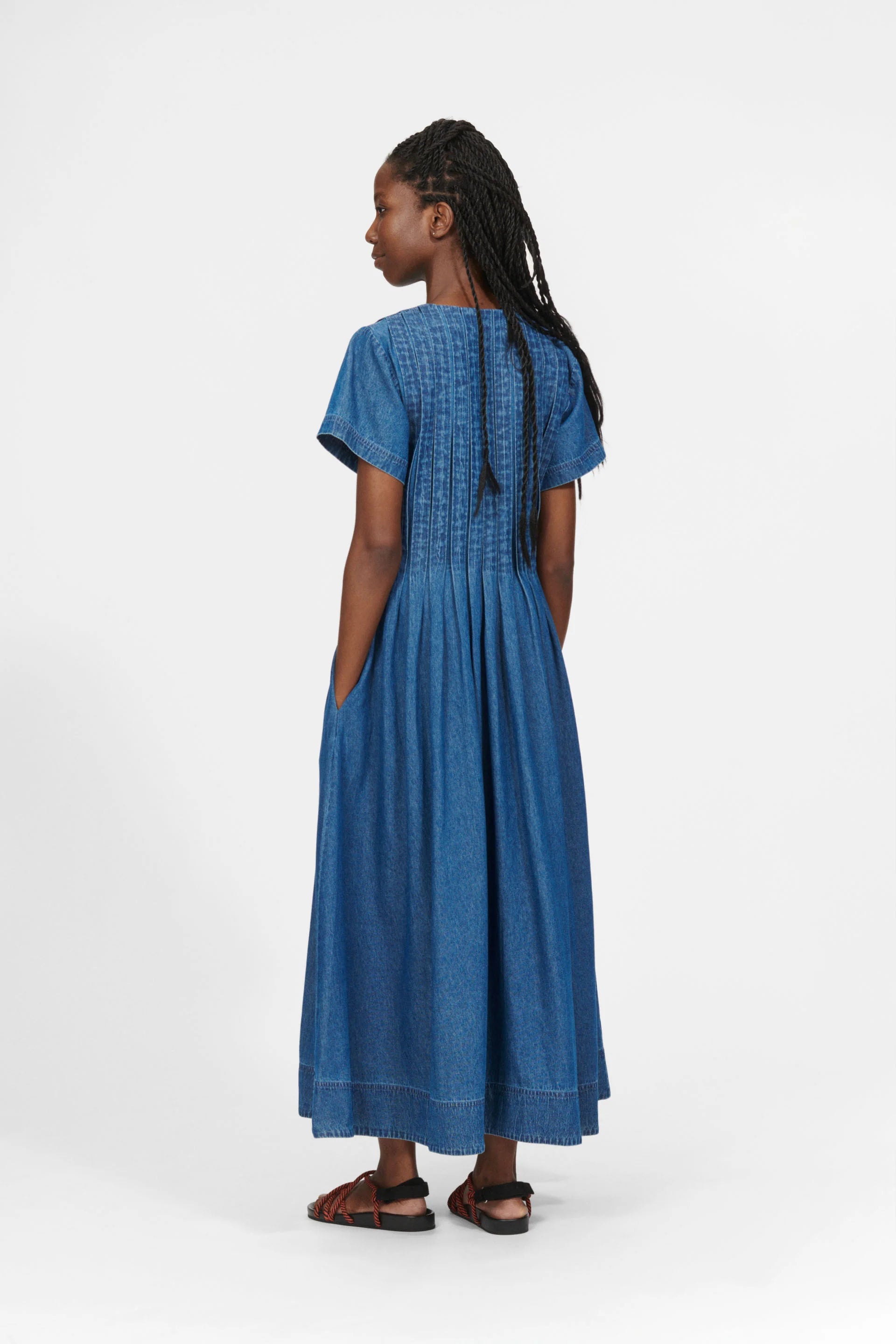 A person with braids wearing the Nué Notes Belize Dress in blue denim, featuring a hidden button closure, paired with sandals, is standing sideways against a plain white background.