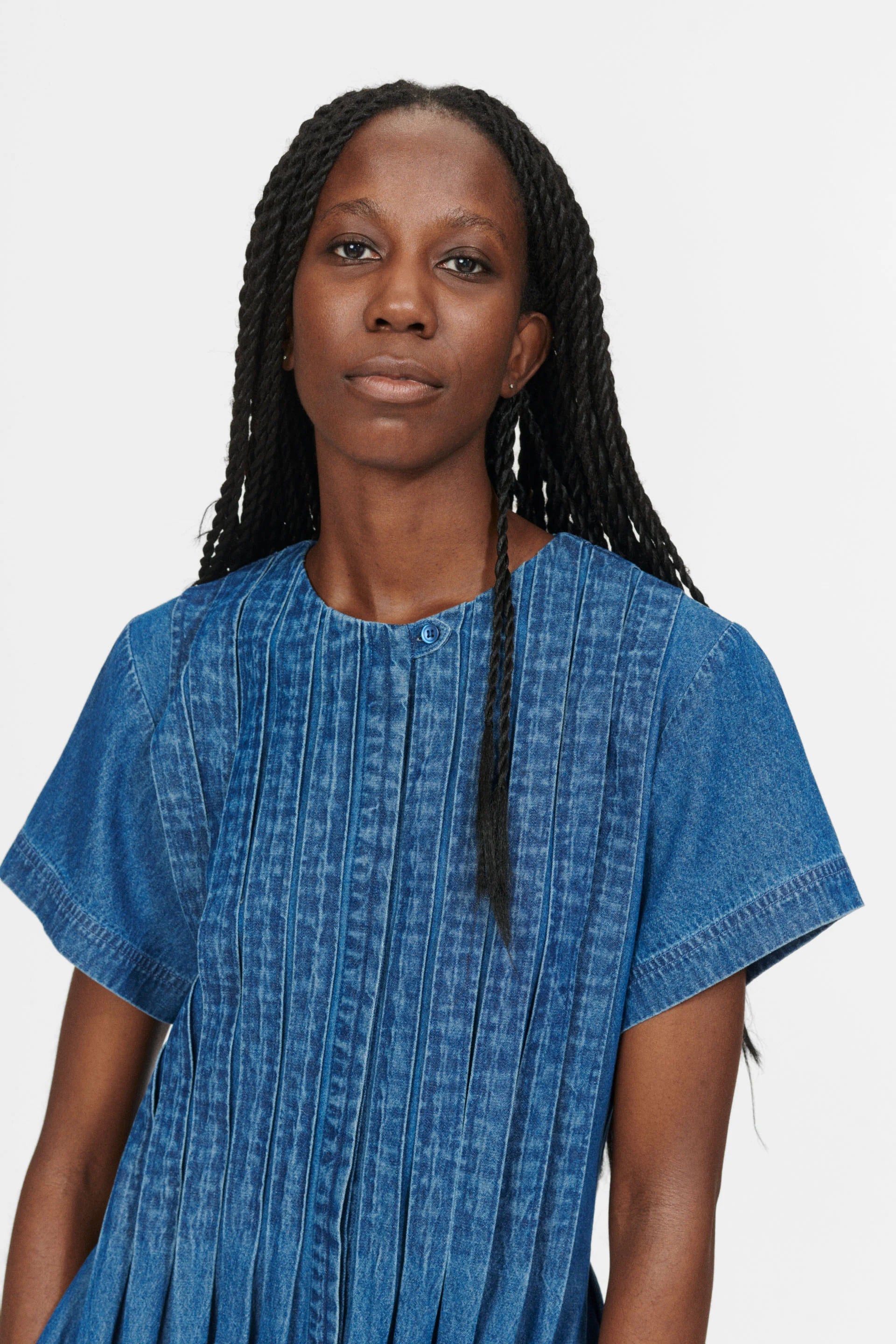 A person with long braided hair is wearing the Belize Dress - Blue Denim by Nué Notes, featuring a short-sleeve style, and is looking at the camera against a plain background.