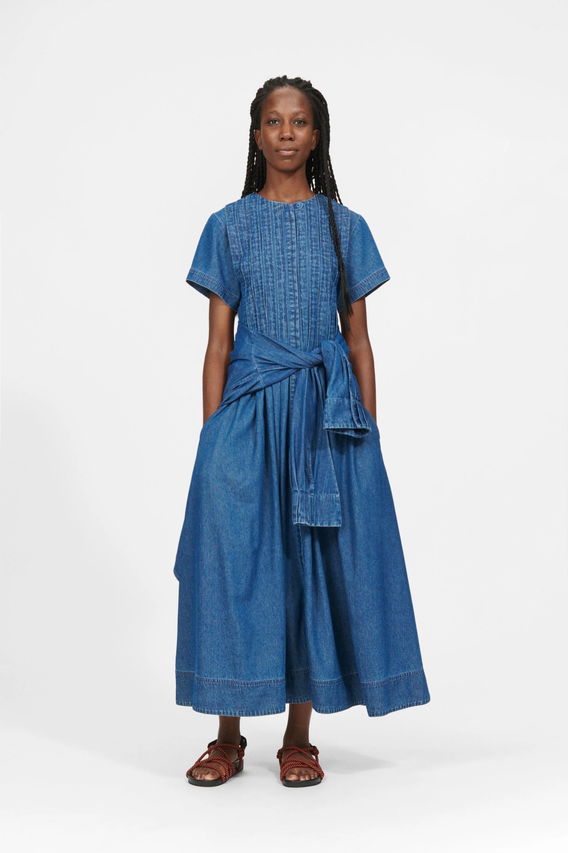 A person stands against a white background wearing the Belize Dress - Blue Denim by Nué Notes. This sleek, stylish midi dress features a tie front and hidden button closure.