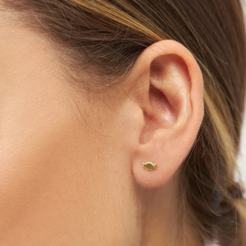 A close-up of a woman's ear wearing a small fish-shaped earring made of gold-plated sterling silver - Lulu Copenhagen Bonbon Single Stud in Gold.