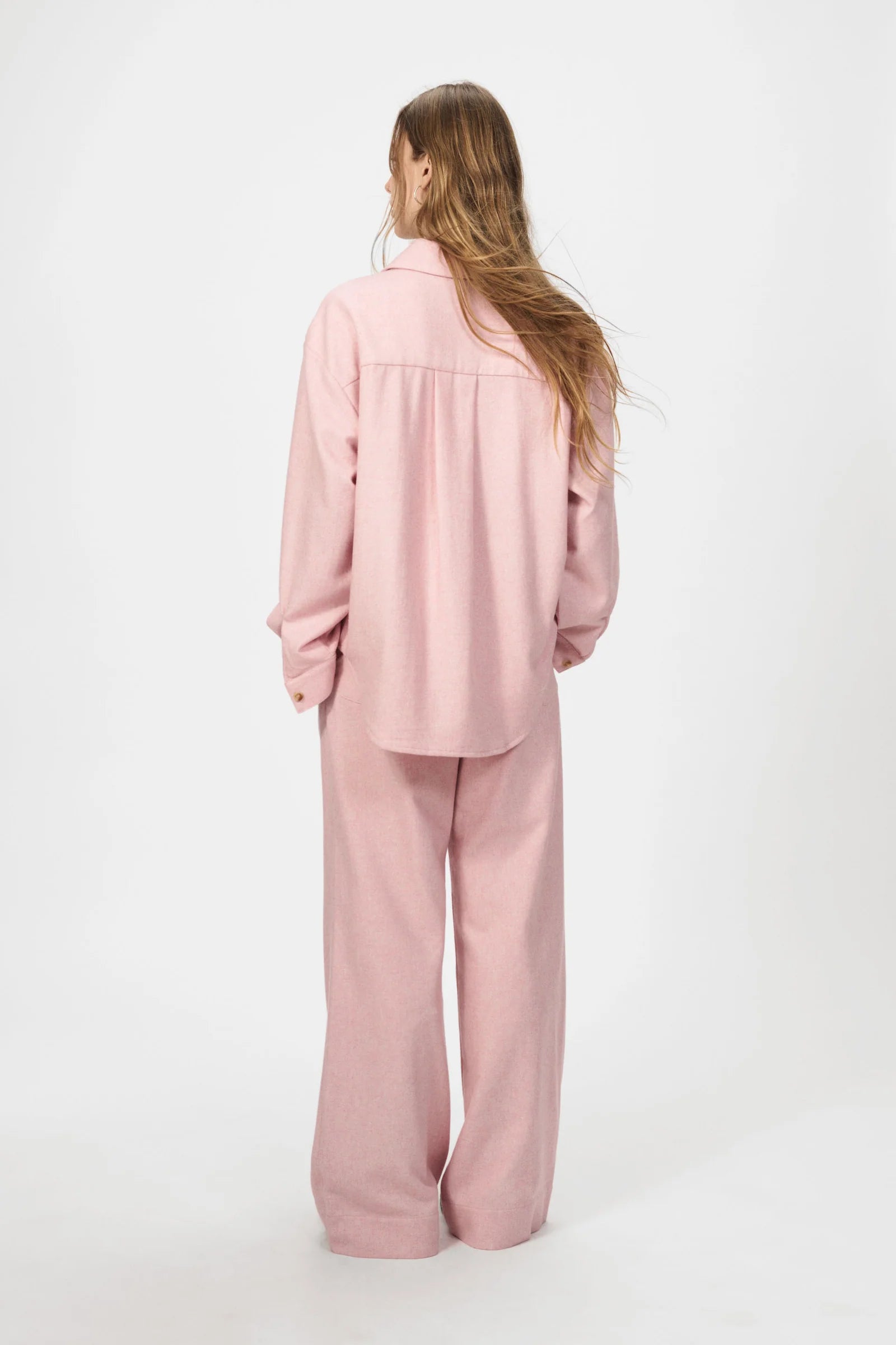 A person stands facing away in the Nué Notes Bonnyrigg Shirt in blush, paired with matching wide-leg pants, against a plain white background.