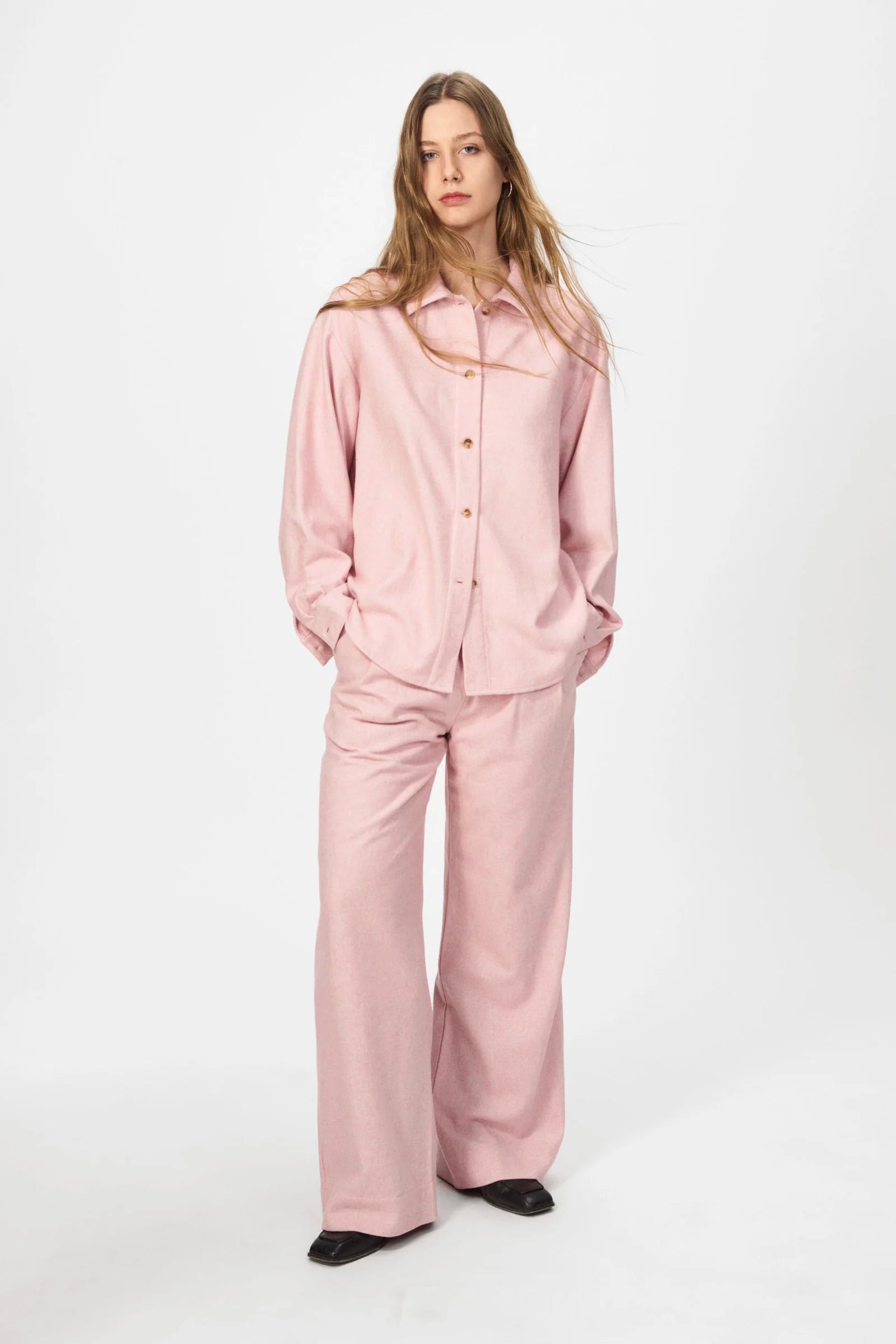 A person stands against a plain white background wearing the oversized Nué Notes Bonnyrigg Shirt in blush melange and matching wide-leg pants, paired with black shoes.