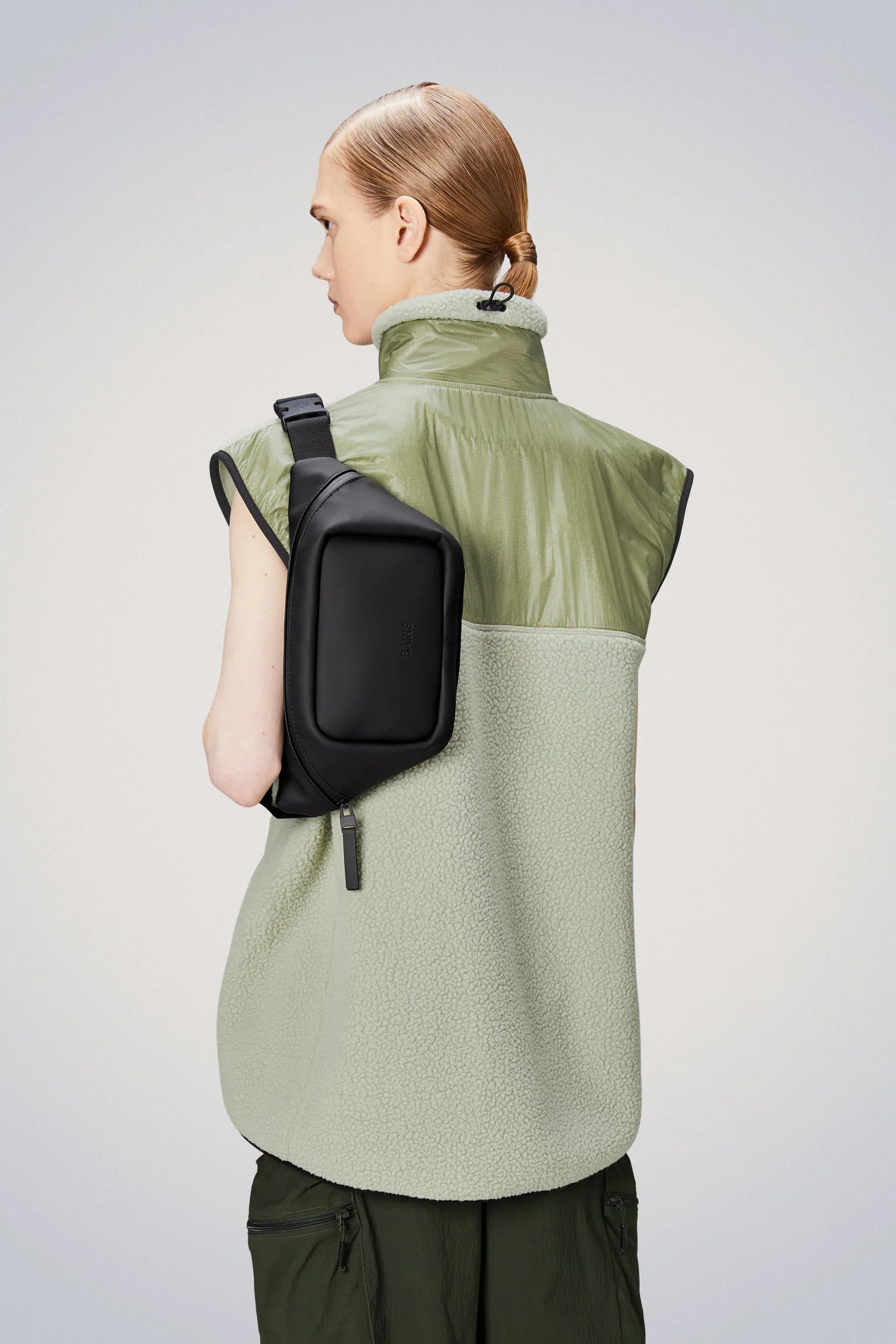 The waterproof Rains Bum Bag Mini, worn on the back of a woman, features an adjustable webbing strap complemented by a green vest.