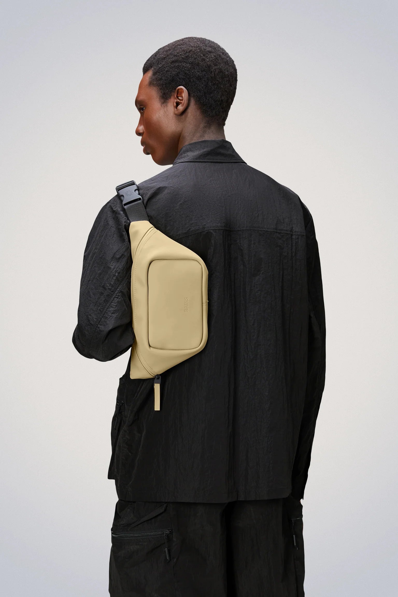A man wearing a black jacket and black pants accessorized with a waterproof Rains Bum Bag Mini featuring an adjustable webbing strap.