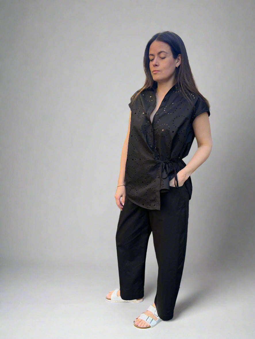 A person wearing MeiMeij's black Barrel Leg Cotton Trousers stylishly stands on a cobblestone street, their ankle-length trousers perfectly complementing the urban backdrop of buildings and parked cars.