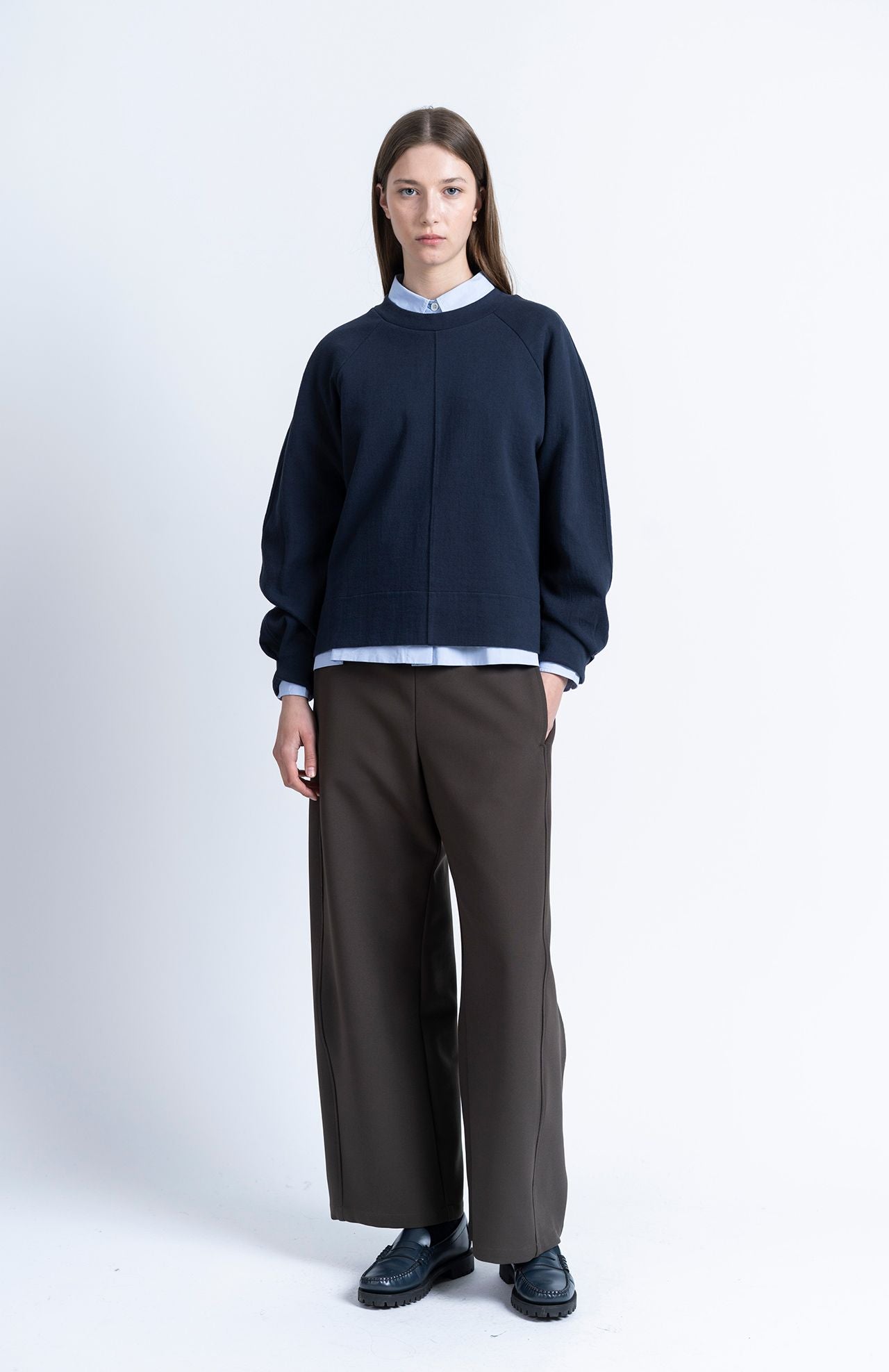 A model wears a relaxed-fit, navy blue Loreak Mendian Celine Sweatshirt over a light blue collared shirt, dark trousers, and black loafers.