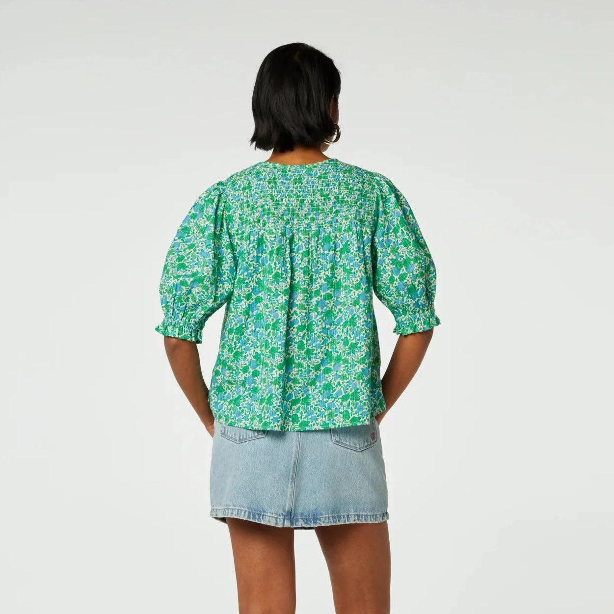 A woman stands facing away from the camera, wearing a June Short Sleeve Organic Top in Green Clueless by Fabienne Chapot and denim shorts against a white background.