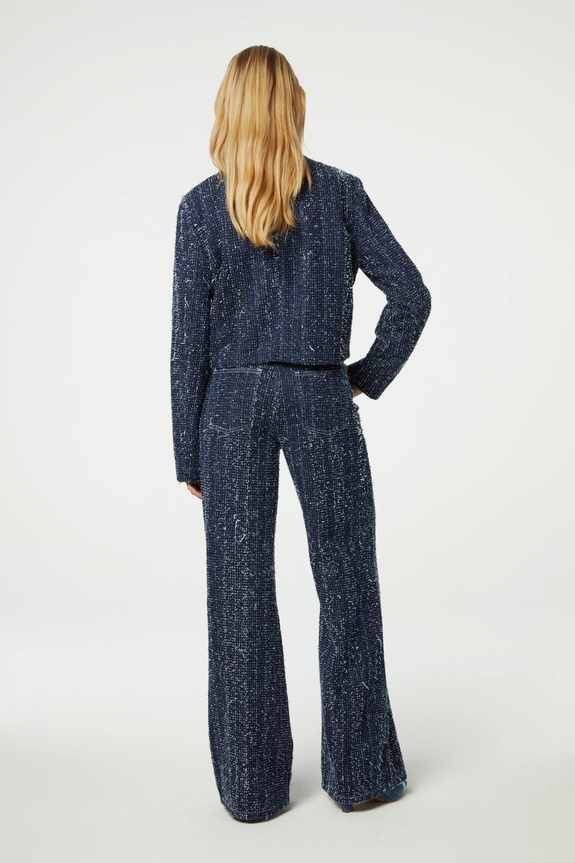 A person with long blonde hair is wearing a navy blue textured pantsuit, complemented by the chic "Davis Blazer" in denim from Fabienne Chapot, standing with their back turned.