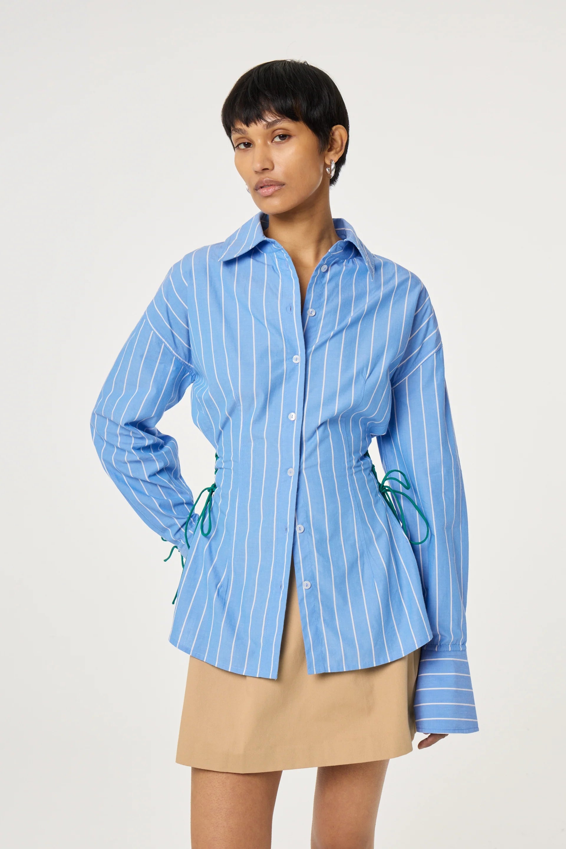 A person wears the Fabienne Chapot Indra Blouse in Marine Blue, featuring blue and white stripes with green side ties, paired with a beige skirt against a plain light background.