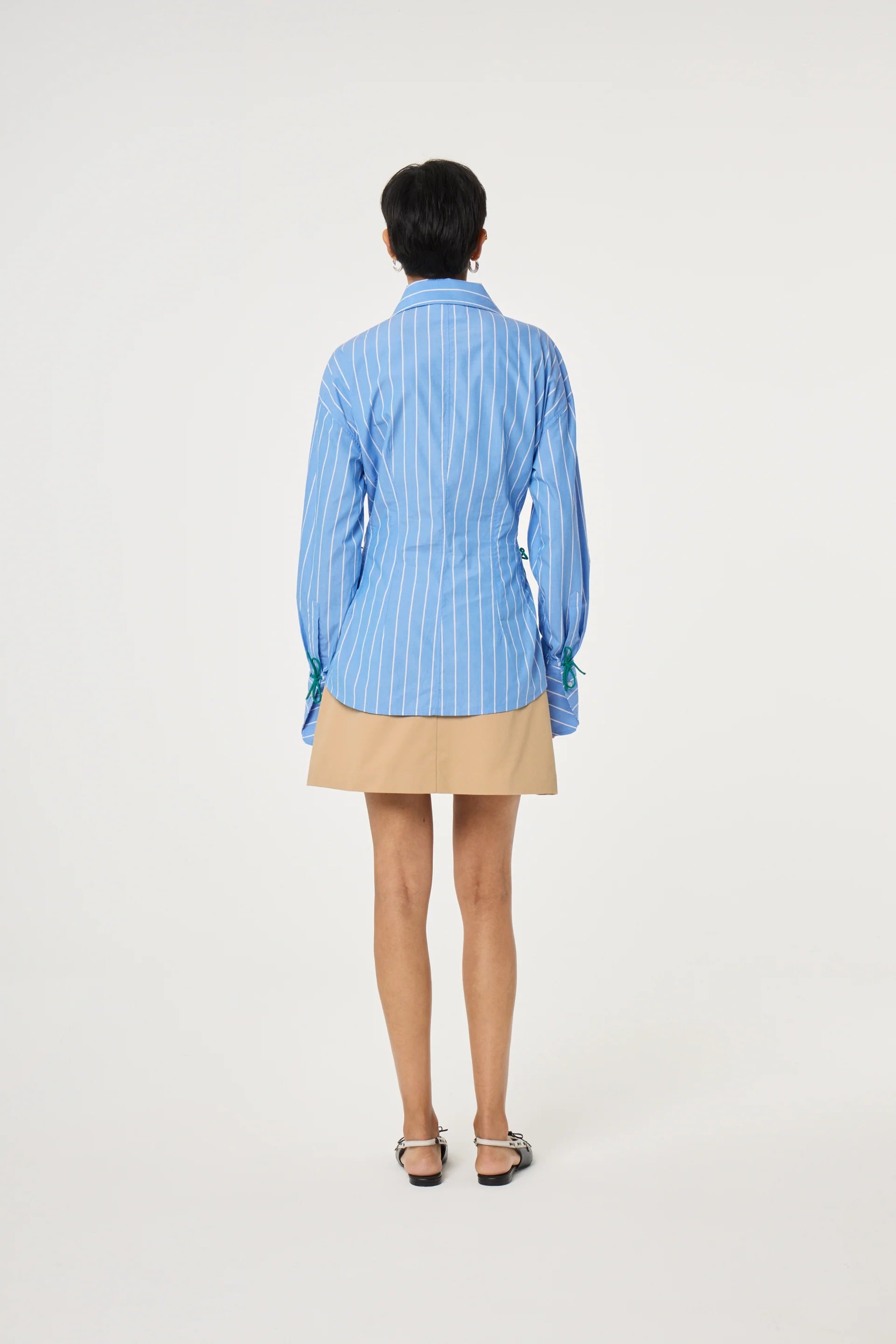 A person faces away, wearing the Fabienne Chapot Indra Blouse in marine blue with blue and white stripes, paired with a beige skirt and flat shoes, set against a plain white background.