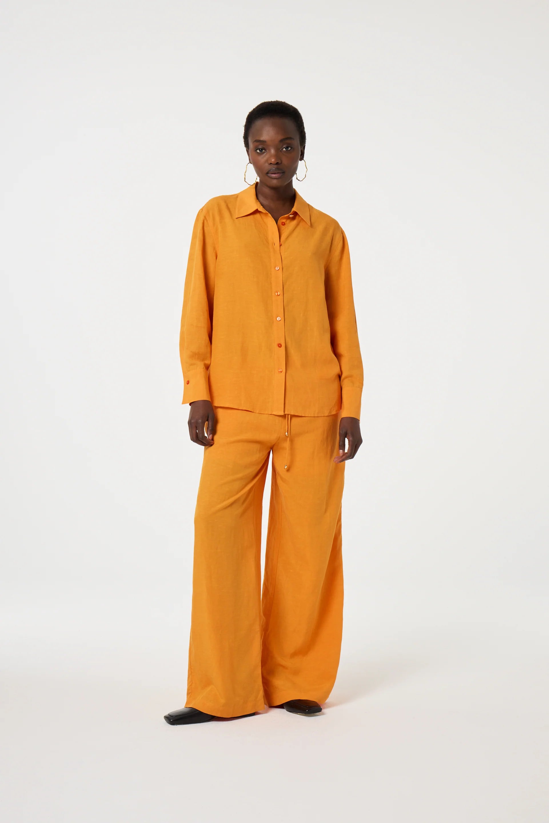 A person stands against a plain white background, wearing the Fabienne Chapot Tony Shirt in Jus d'Orange with matching wide-leg pants and black shoes.