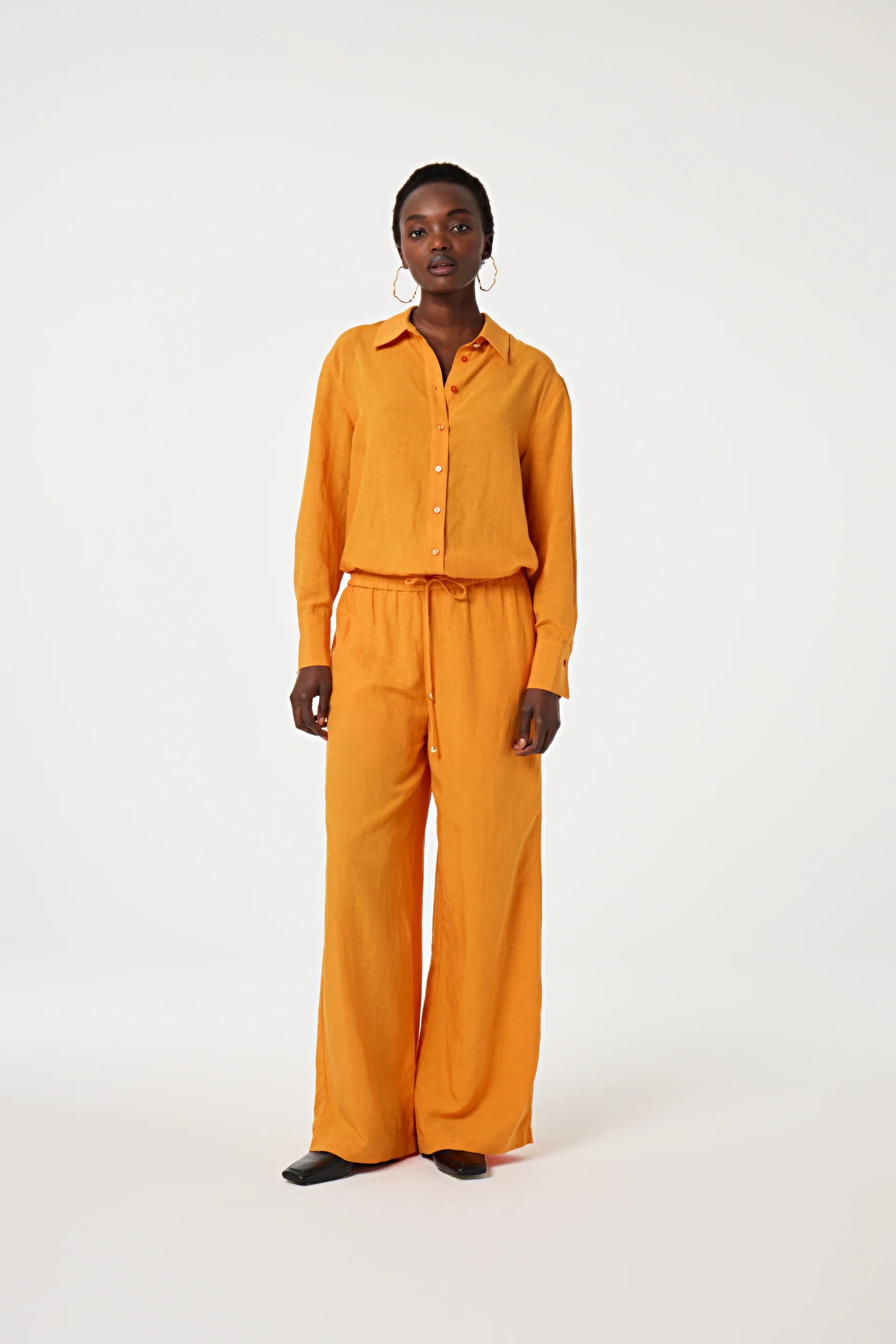 A person stands facing forward in the Fabienne Chapot Tony Shirt - Jus d'Orange, paired with matching wide-leg pants and black shoes, set against a plain white background.