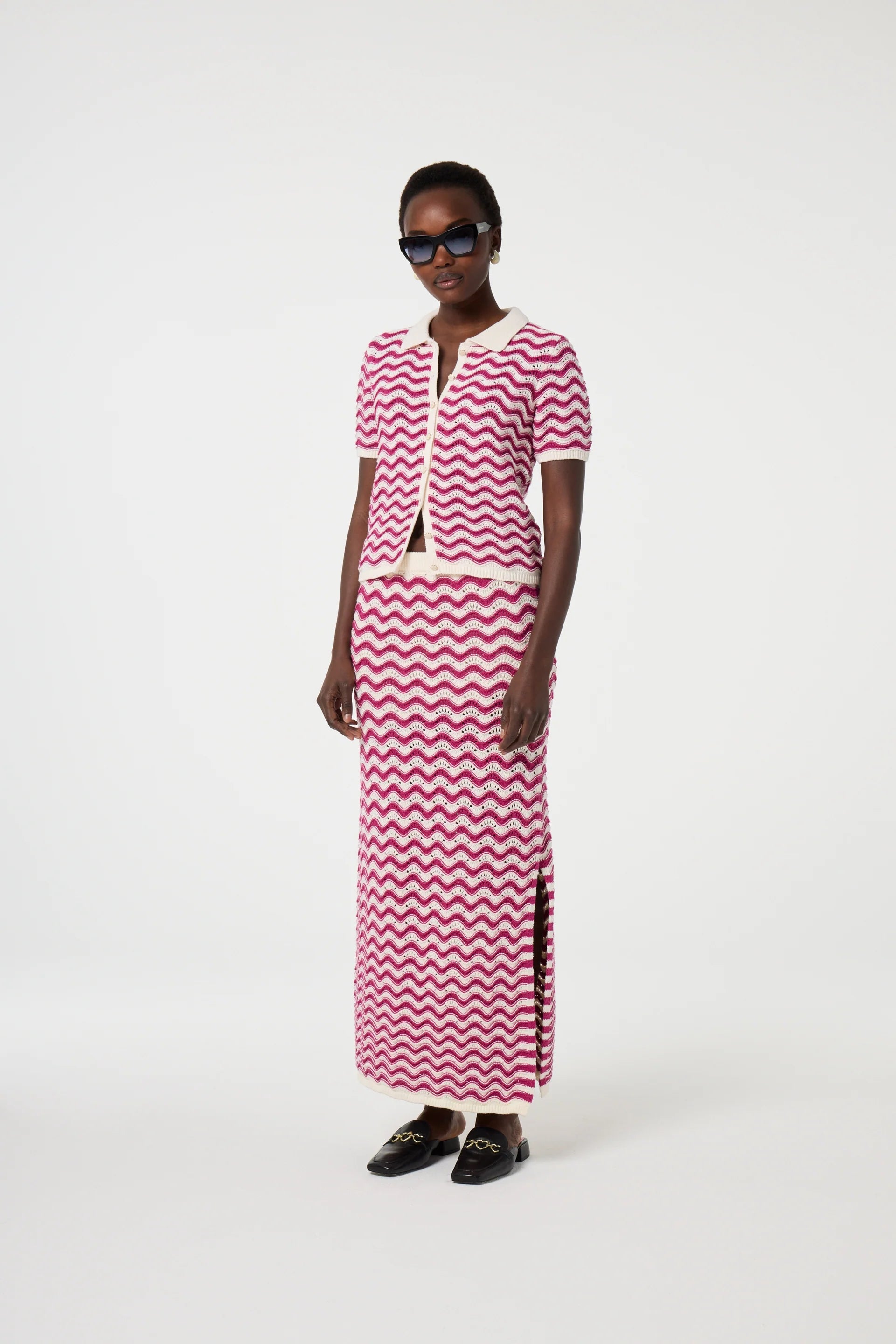 A person in Fabienne Chapot’s Caro Crochet Cardigan – Pink Craze, paired with a matching skirt and sunglasses, stands against a plain white background.