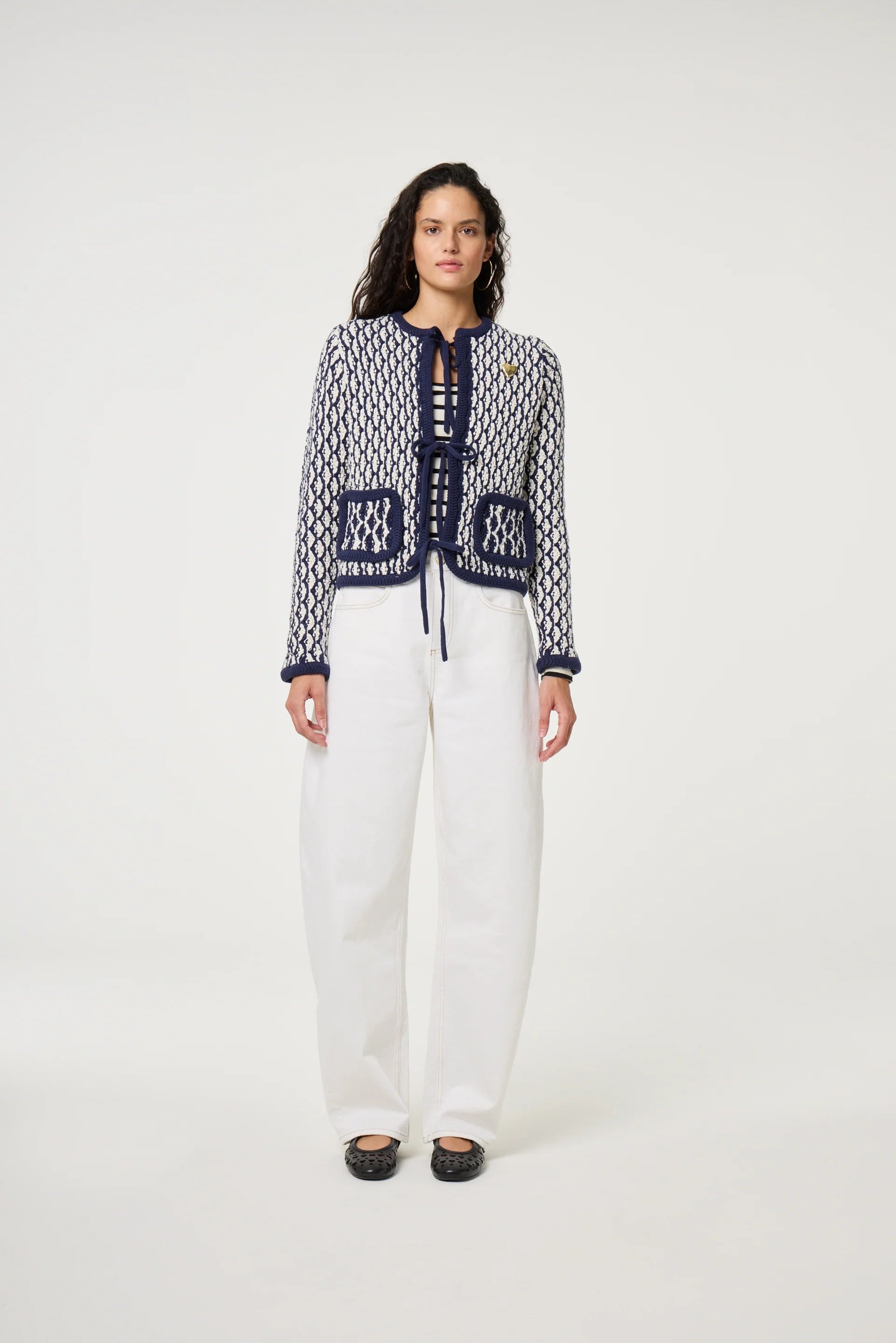 A woman stands against a plain background wearing Fabienne Chapot’s Betty Barrel Jeans in white, a patterned navy and white organic cotton cardigan, and black shoes.