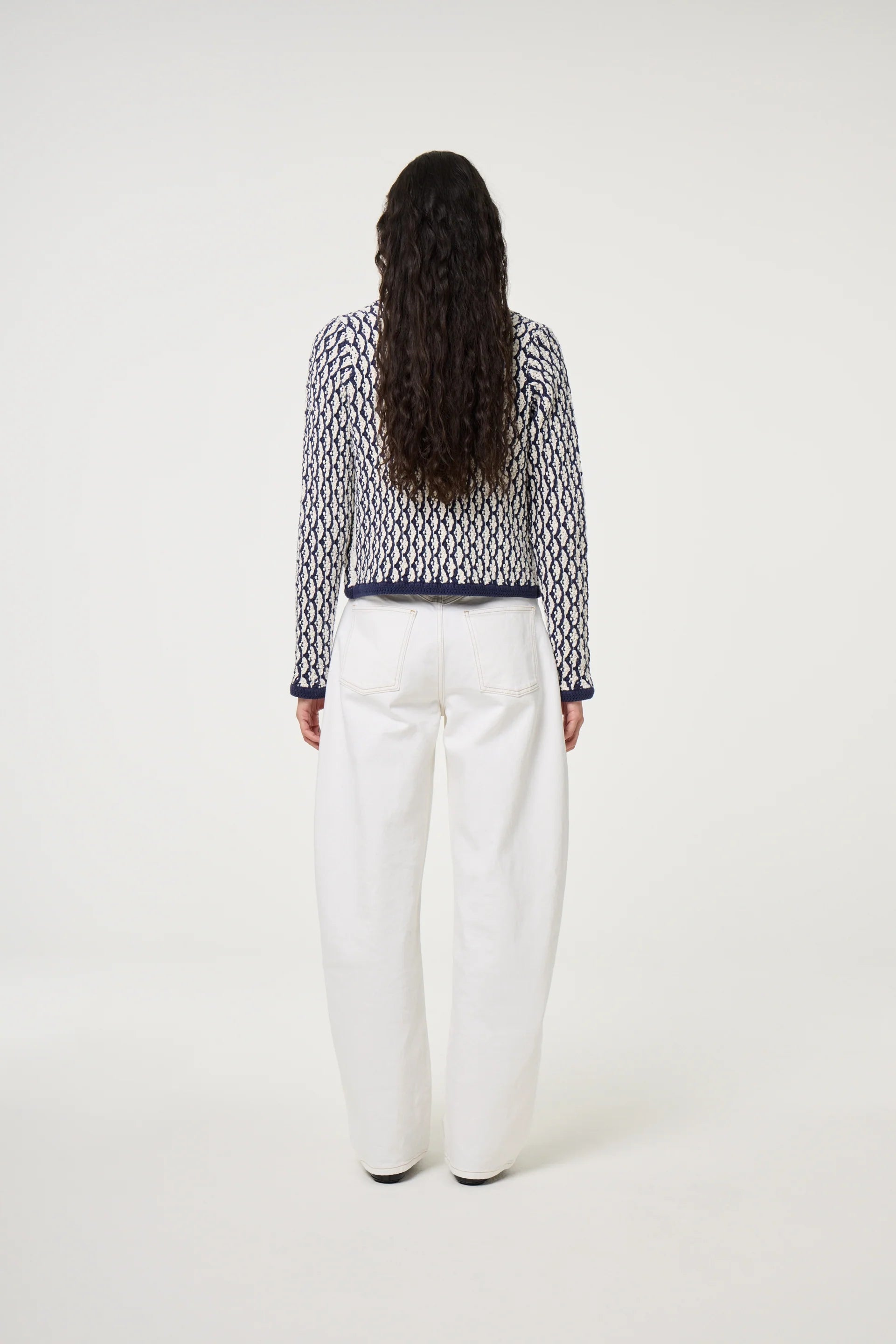 A person with long curly hair stands facing away, wearing a navy patterned sweater and Fabienne Chapot’s Betty Barrel Jeans in white, featuring an organic cotton relaxed silhouette, against a plain light background.