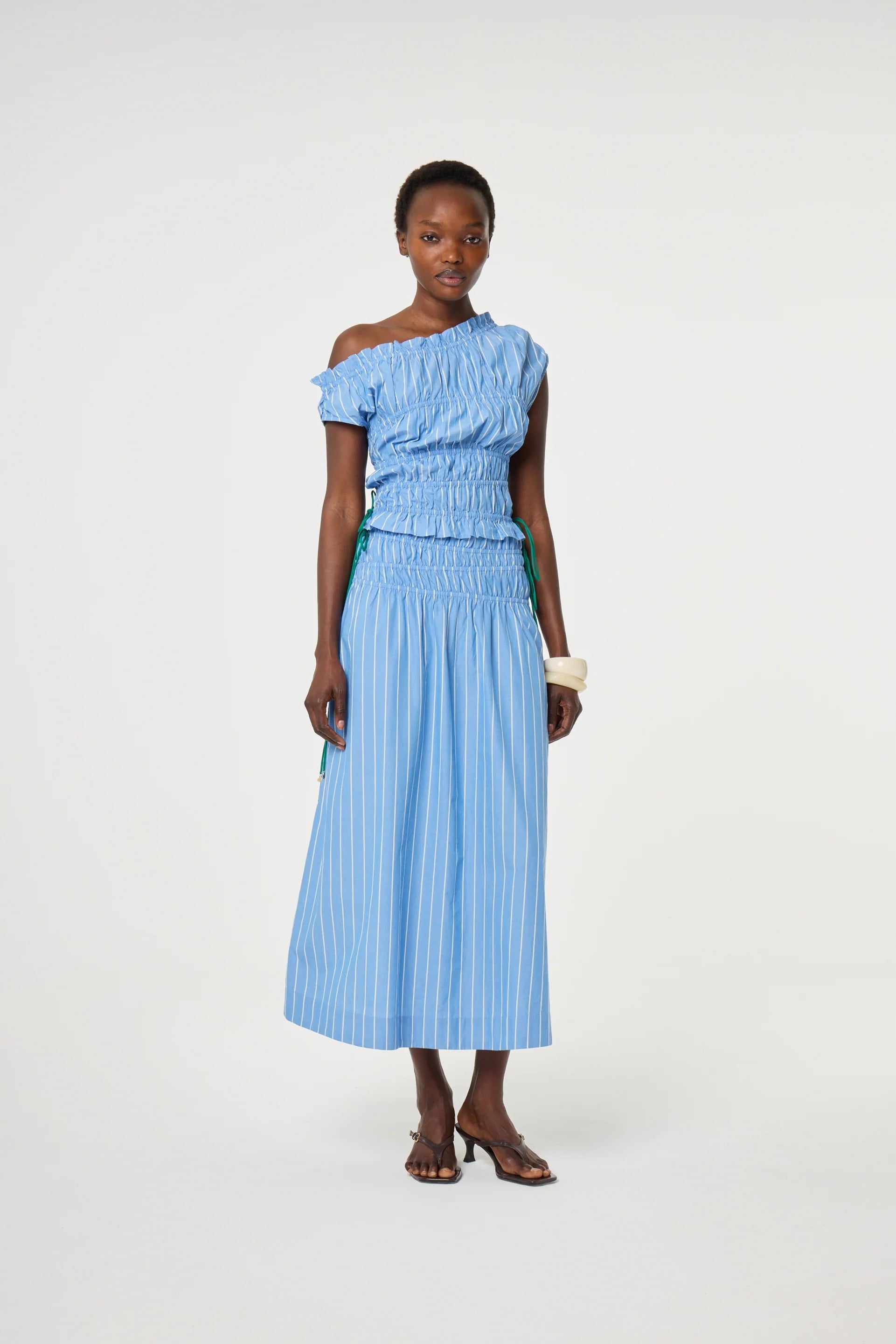 A woman stands against a plain white background wearing the Fabienne Chapot John Ruched Top in Marine Blue, styled with a light blue striped midi skirt, dark sandals, and a small clutch bag.