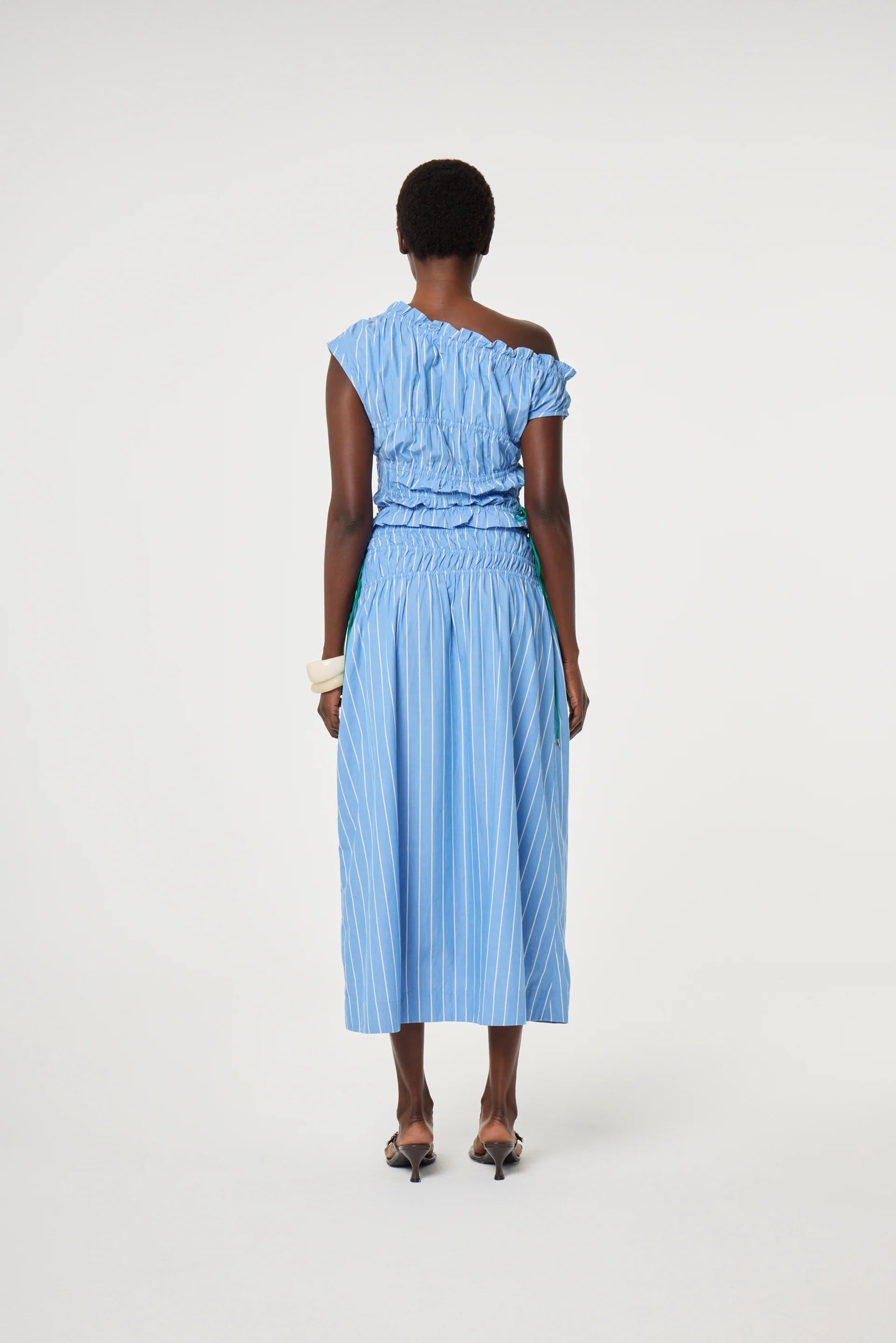 A person stands with their back to the camera, wearing the Fabienne Chapot John Ruched Top in Marine Blue, paired with a matching blue and white striped midi skirt, high heels, and a white bangle.