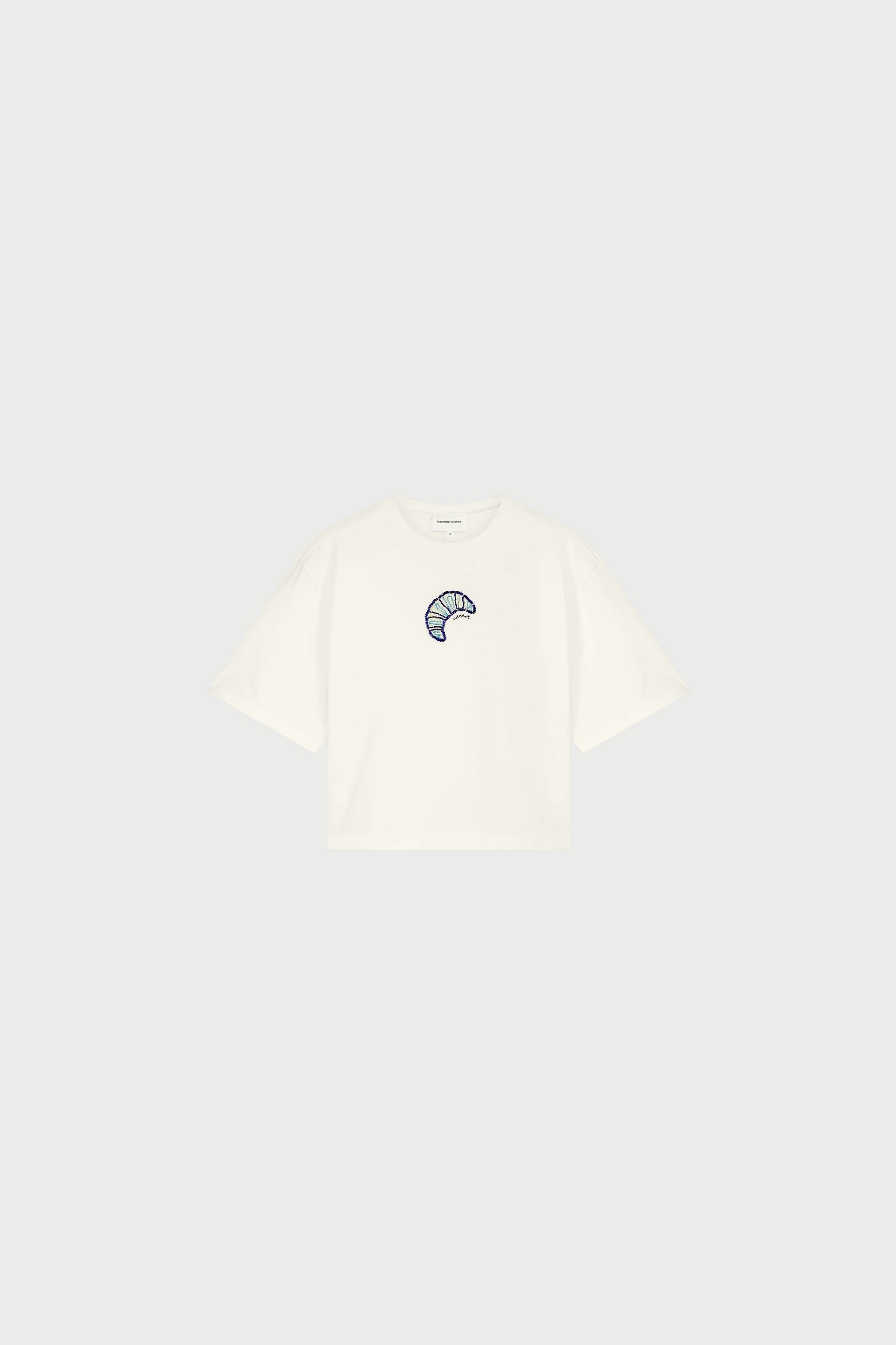 The Benja Tshirt - Cosy White by Fabienne Chapot is a relaxed-fit organic cotton T-shirt with a small, colorful croissant print on the center chest, shown here against a light gray background.