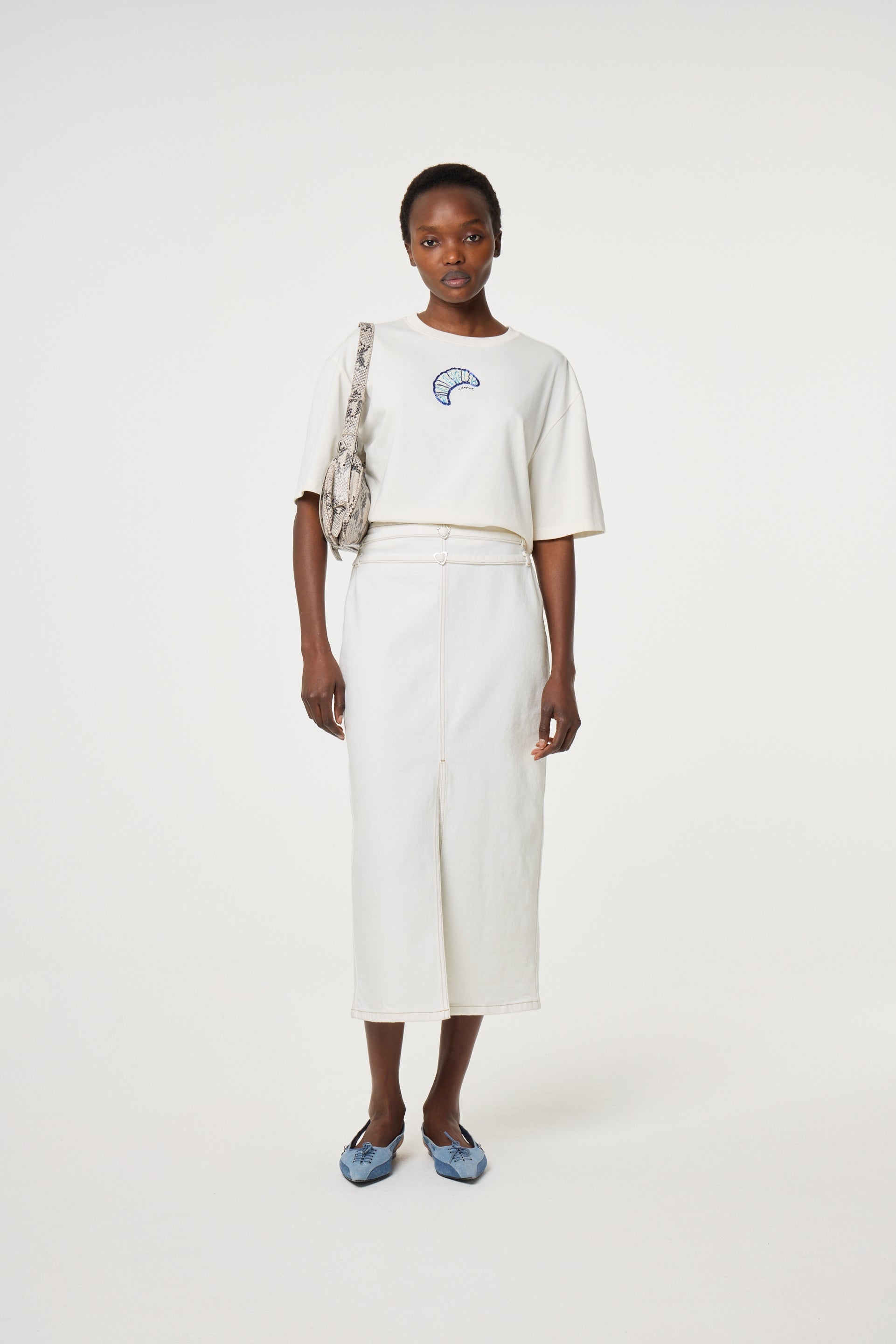 A woman faces forward against a plain white background, wearing the Fabienne Chapot Benja Tshirt in Cosy White—a relaxed-fit tee with a small blue graphic—paired with a white midi skirt, blue flats, and a gray patterned shoulder bag.