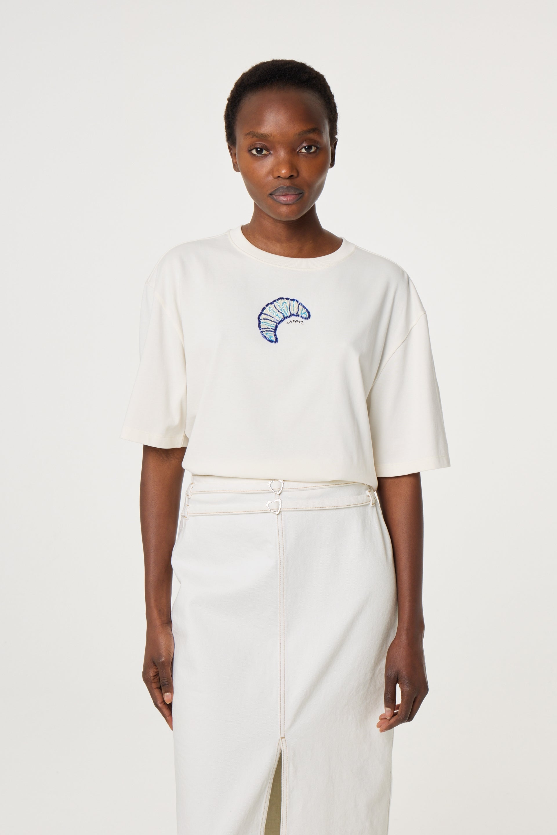 A person is shown facing forward wearing the Fabienne Chapot Benja Tshirt in Cosy White, a relaxed-fit short-sleeve shirt featuring a blue and purple embroidered design, paired with a white front-slit skirt.