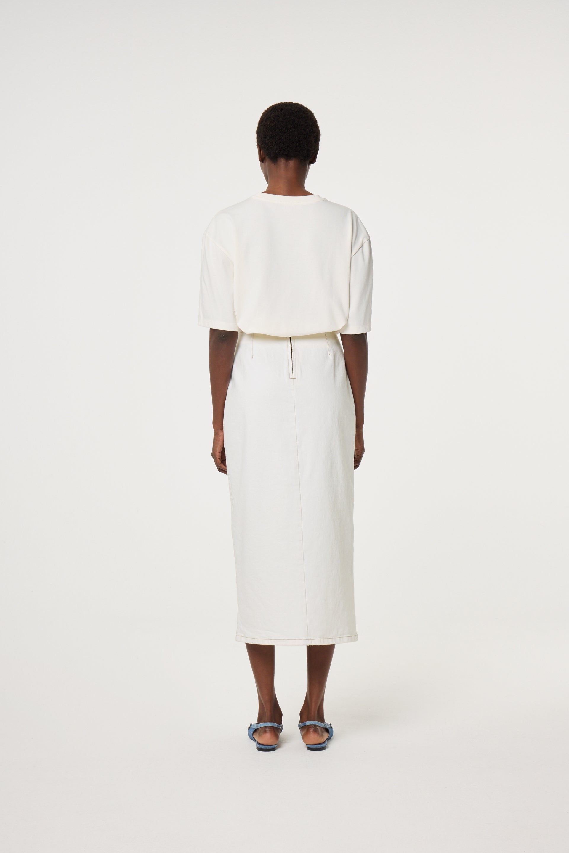 A person stands facing away from the camera, wearing the Fabienne Chapot Benja Tshirt in Cosy White, a white midi skirt, and blue sandals against a plain white background.