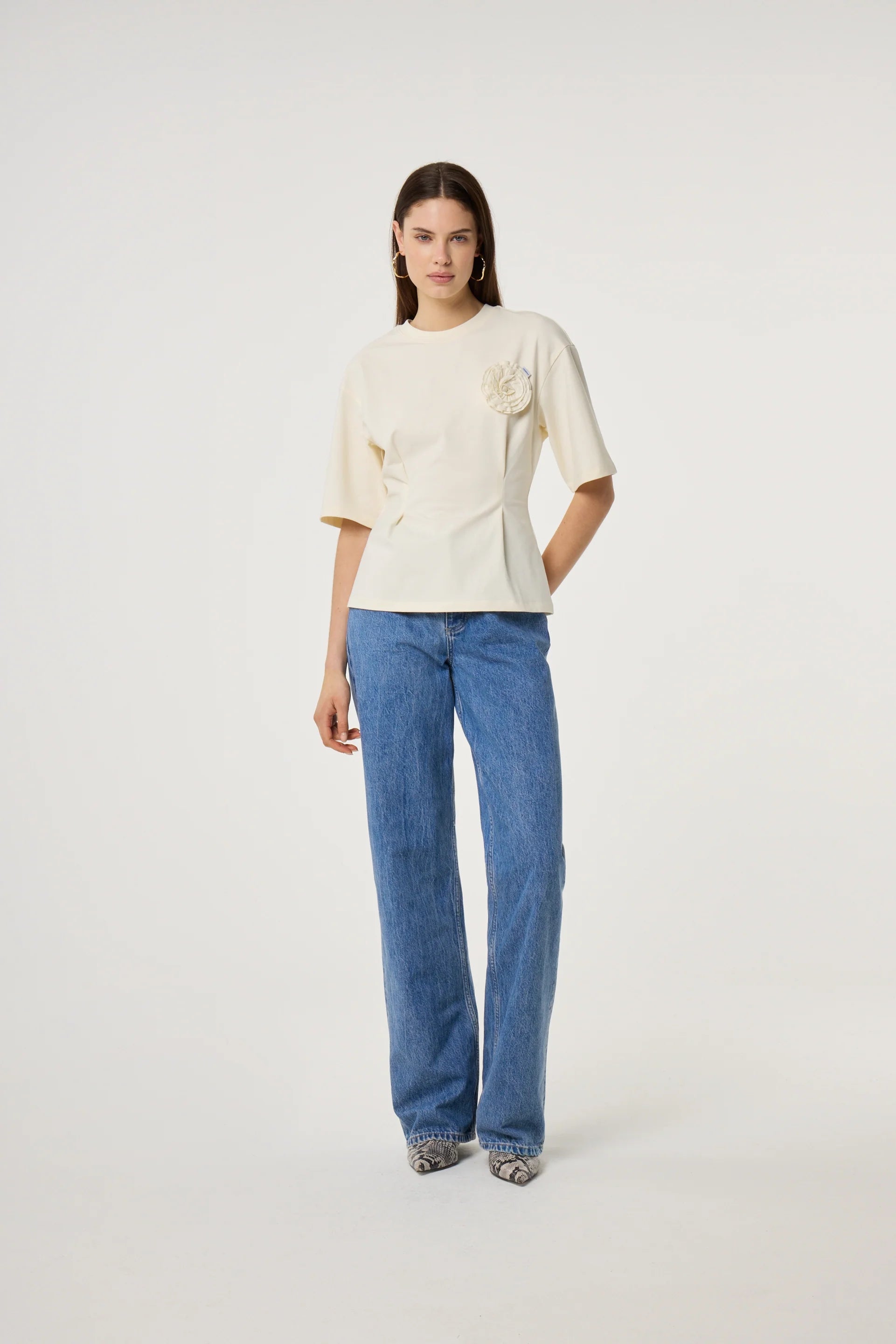 A woman stands against a white background wearing the Fabienne Chapot Asta T-shirt in Cosy White, a cream organic cotton tee with a floral accent, paired with high-waisted blue wide-leg jeans.