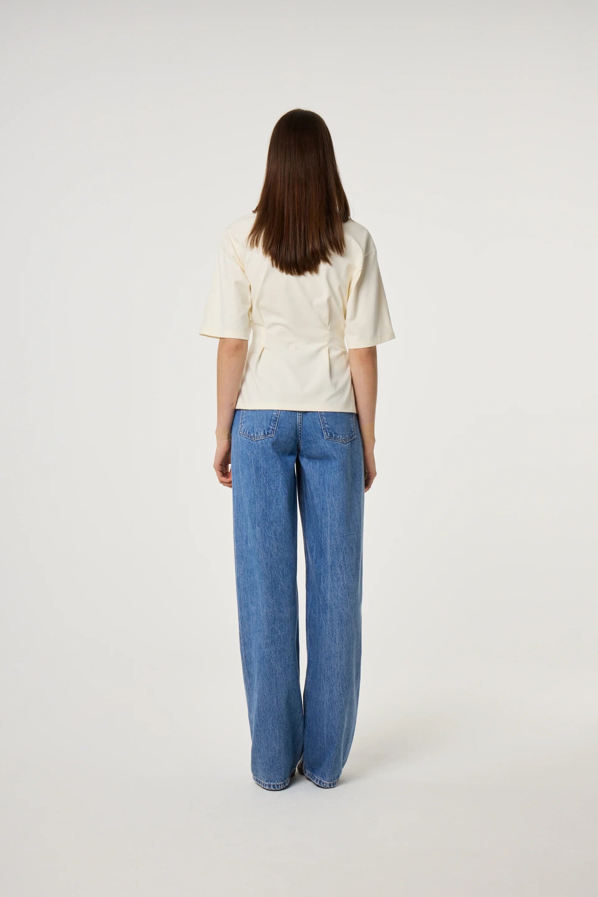 A person with straight brown hair, wearing the Fabienne Chapot Asta T-shirt in Cosy White and blue wide-leg jeans, stands facing away against a plain white background.