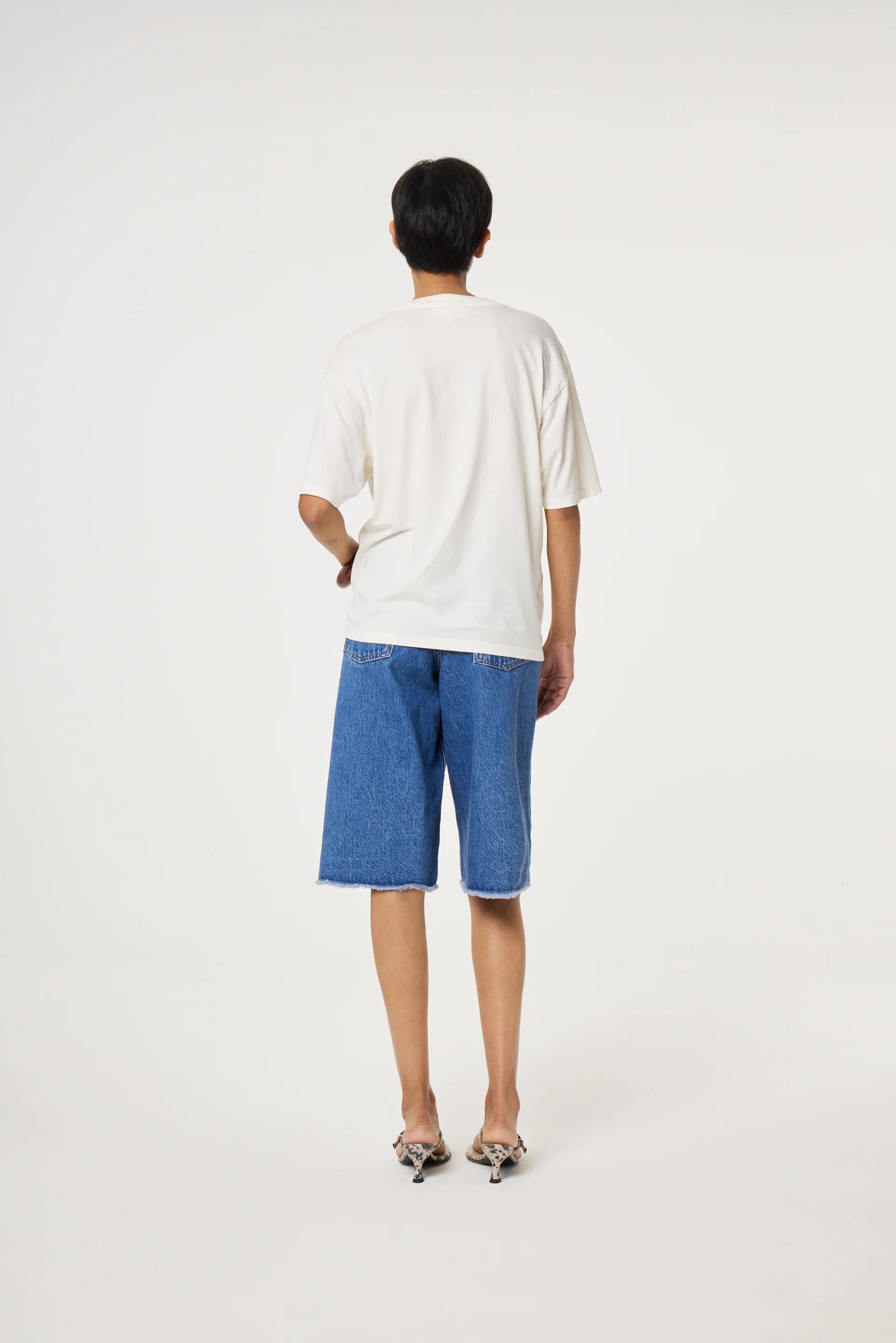A person stands with their back to the camera, wearing the Fabienne Chapot Steve T-shirt in Cosy White, blue knee-length denim shorts, and flat shoes against a plain white background.