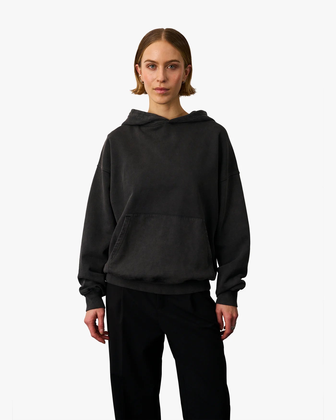 A person stands against a plain background wearing the Organic Oversized Hood in Faded Black from Colorful Standard and black pants.