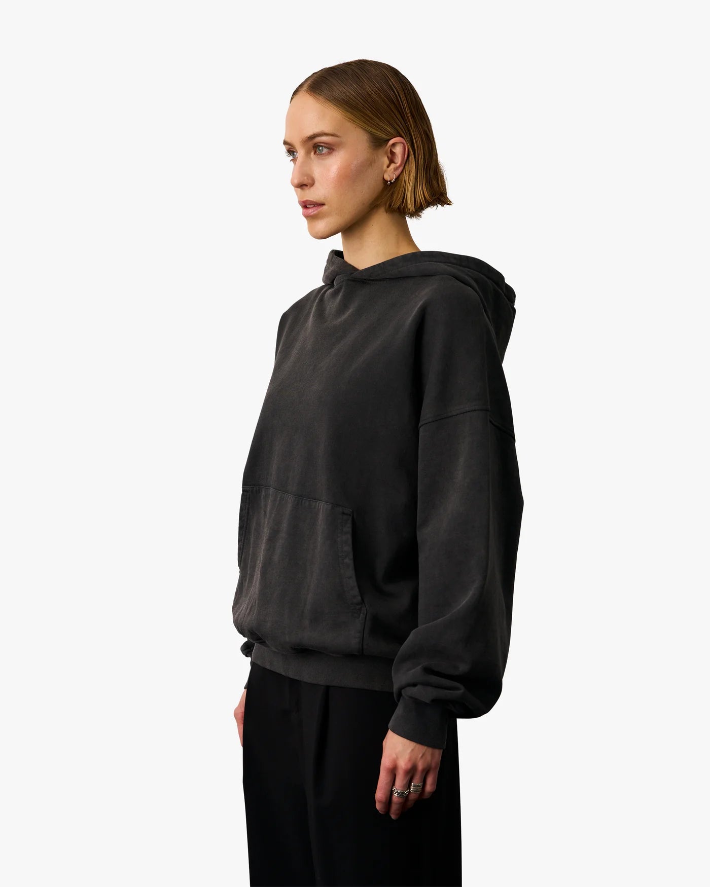 A person with short hair is wearing a Colorful Standard Organic Oversized Hood in Faded Black and black pants, looking to the left. The background is plain white.