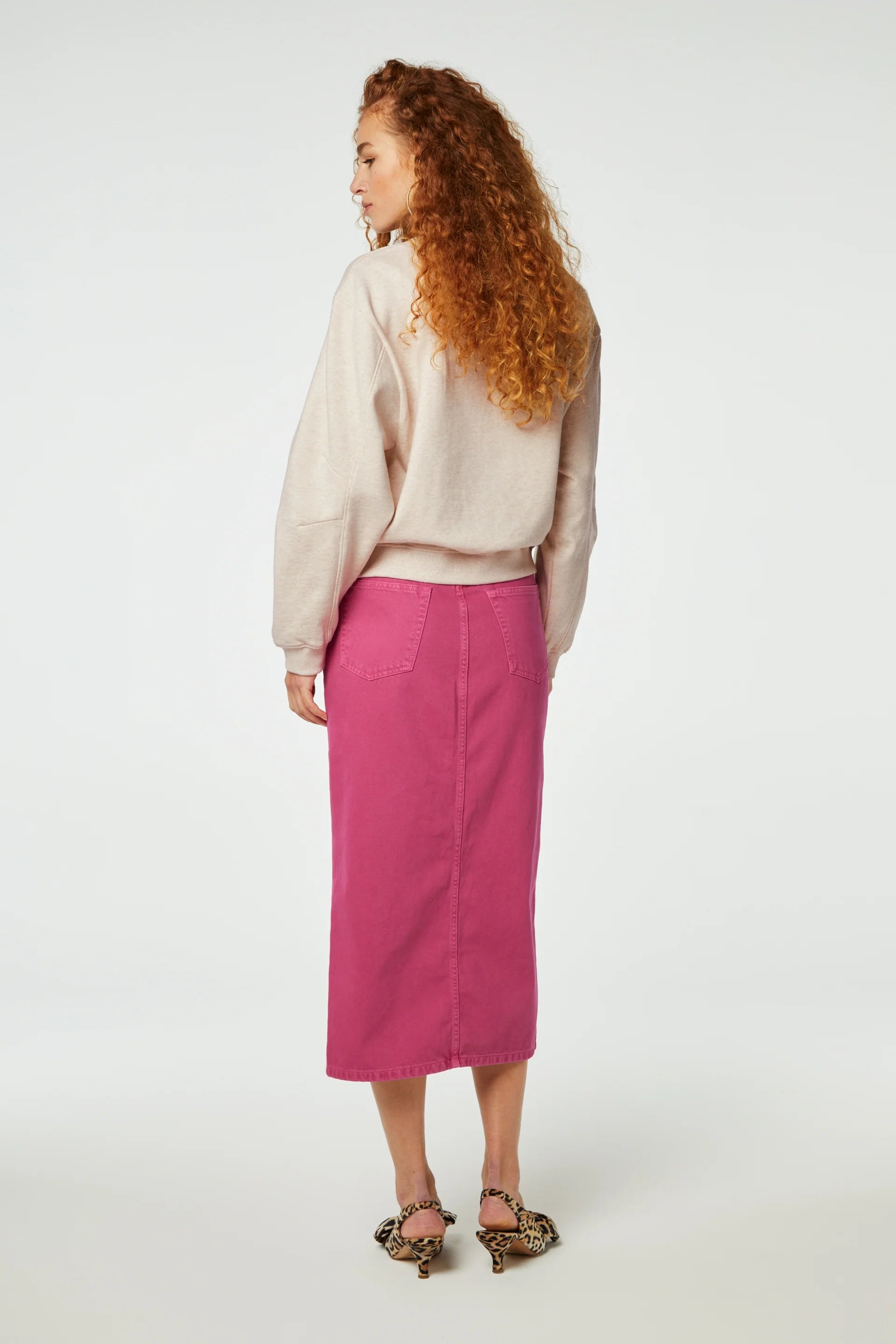 Woman standing with her back to the camera wearing a beige sweatshirt and a Carlyne skirt in hot pink from Fabienne Chapot, paired with patterned heeled sandals.