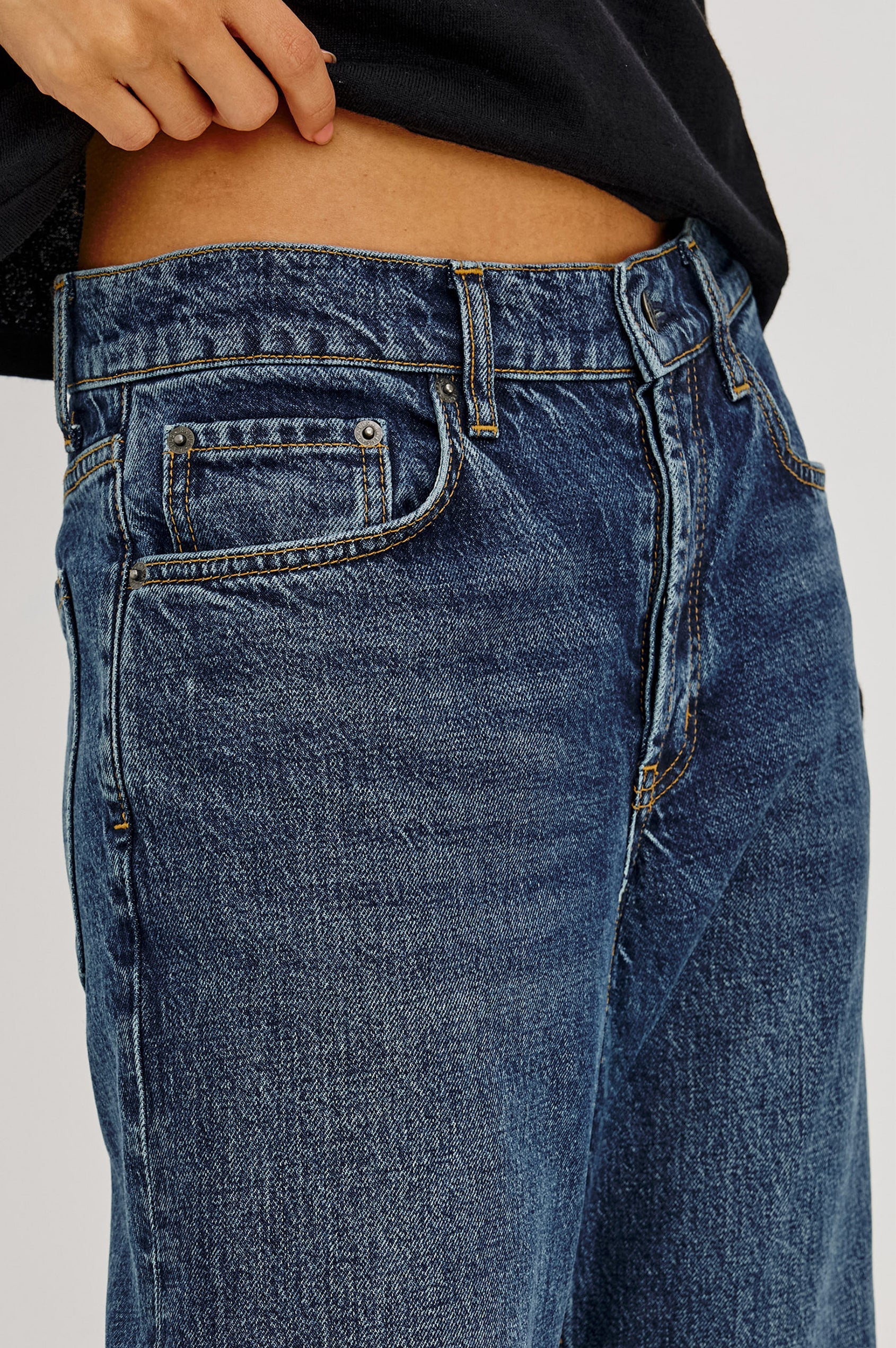 Close-up of a person wearing The Del Mar - Upstream low-rise vintage jeans by Rails and a black shirt, with one hand resting on the waistband.