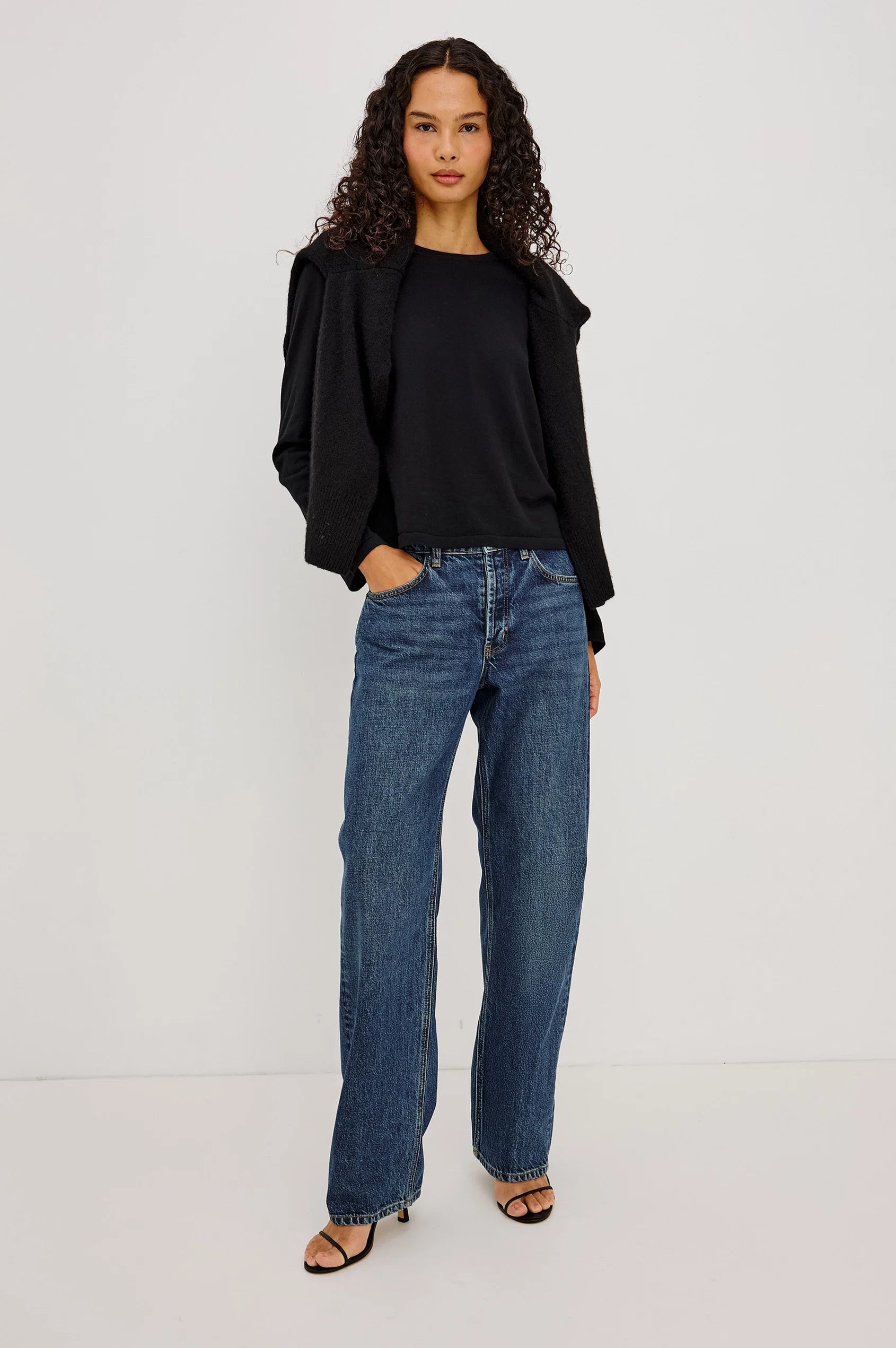 A person stands against a plain white background with hands in their pockets, wearing Rails’ The Del Mar - Upstream relaxed wide-leg jeans, a black shirt, a black sweater draped over their shoulders, and black heeled sandals.