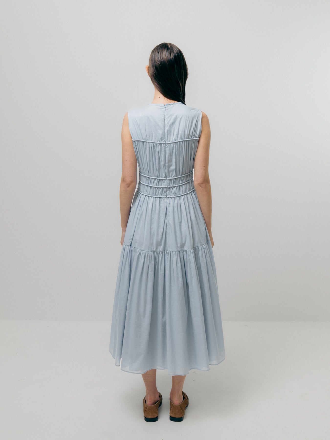 A woman with long dark hair stands facing away against a light background, dressed in the Emin and Paul Triple Gathered Dress in Sky Blue, featuring a pure cotton fabric and an elasticated waist, paired with sandals.