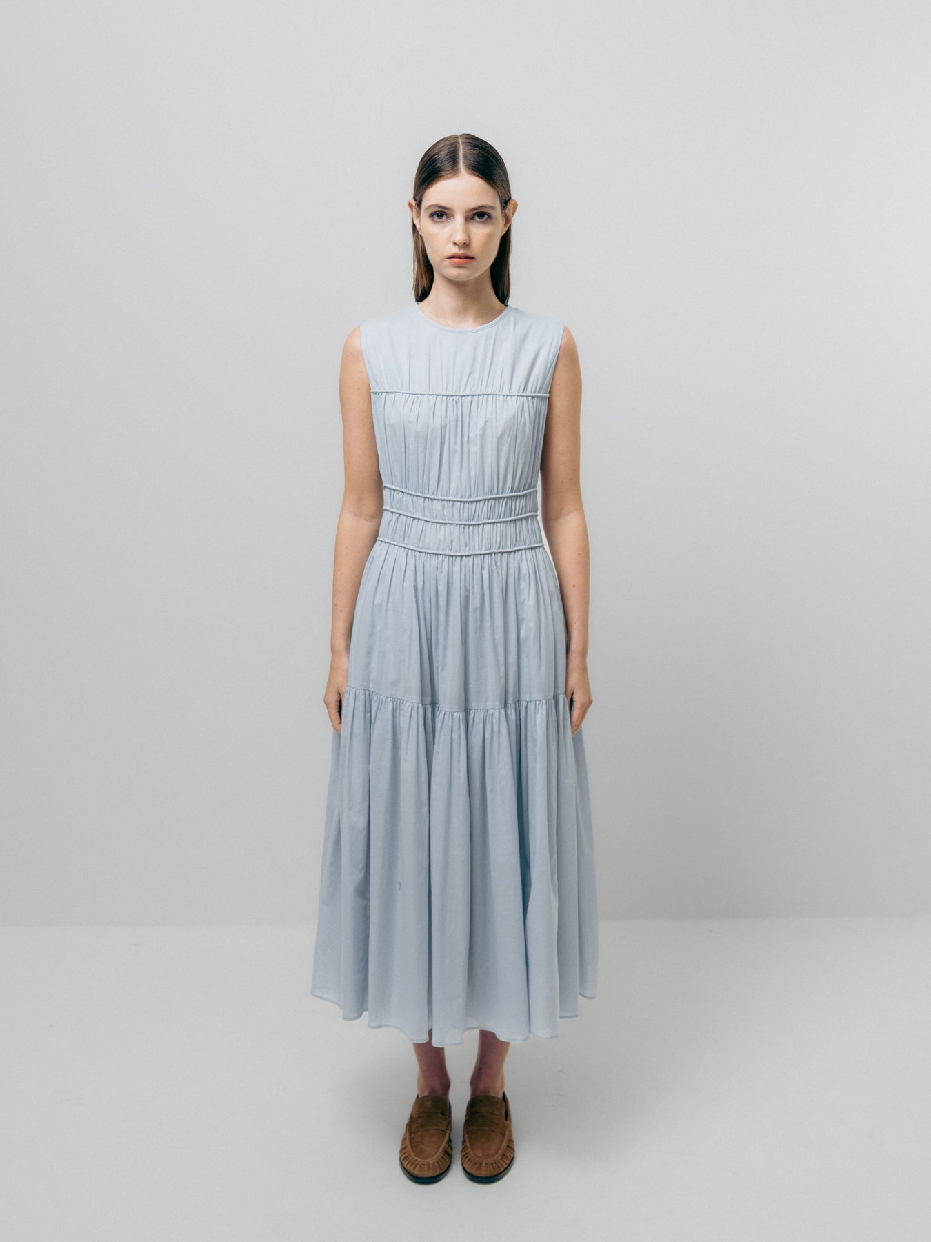 A person wearing the Triple Gathered Dress - Sky Blue by Emin and Paul, featuring a sleeveless pure cotton design with an elasticated waist, stands against a plain background, looking directly at the camera.