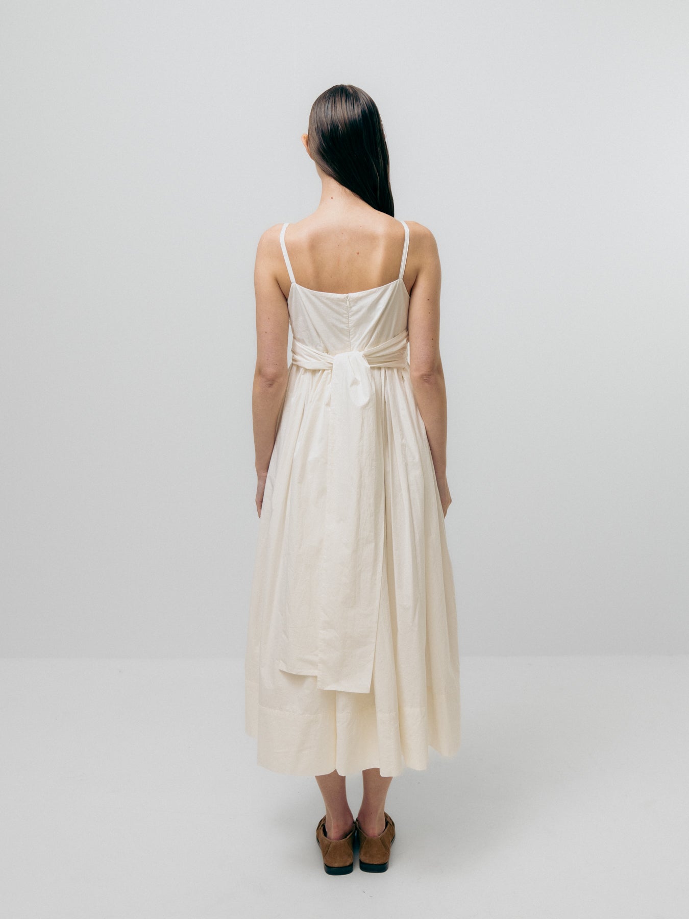 In a minimalist, light-colored space, a person with long dark hair faces away while wearing the Tie Front Dress - White by Emin and Paul.