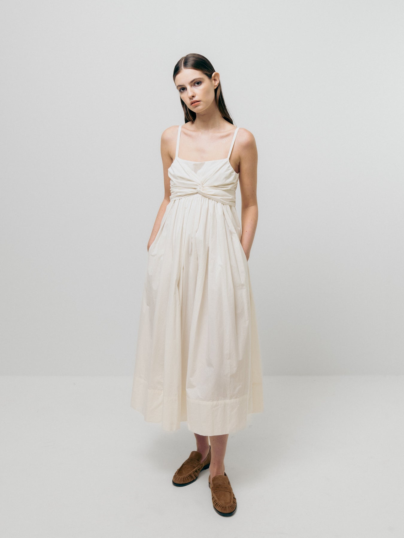 A person wearing a Tie Front Dress by Emin and Paul, featuring a sleeveless design and flowy silhouette in cream-white, pairs it with brown shoes against a plain white background.