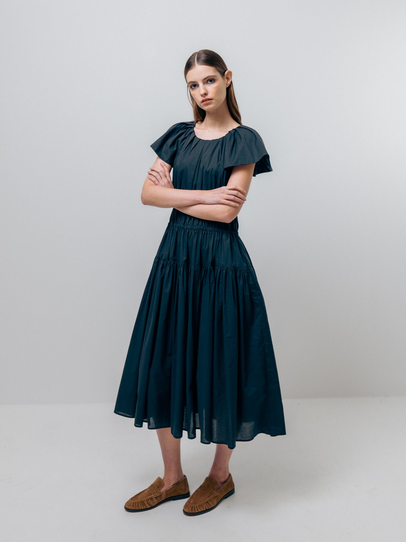 A woman stands against a plain background, wearing an Emin and Paul Wide Shoulder Dress in navy with flutter sleeves and brown shoes, her arms confidently crossed.