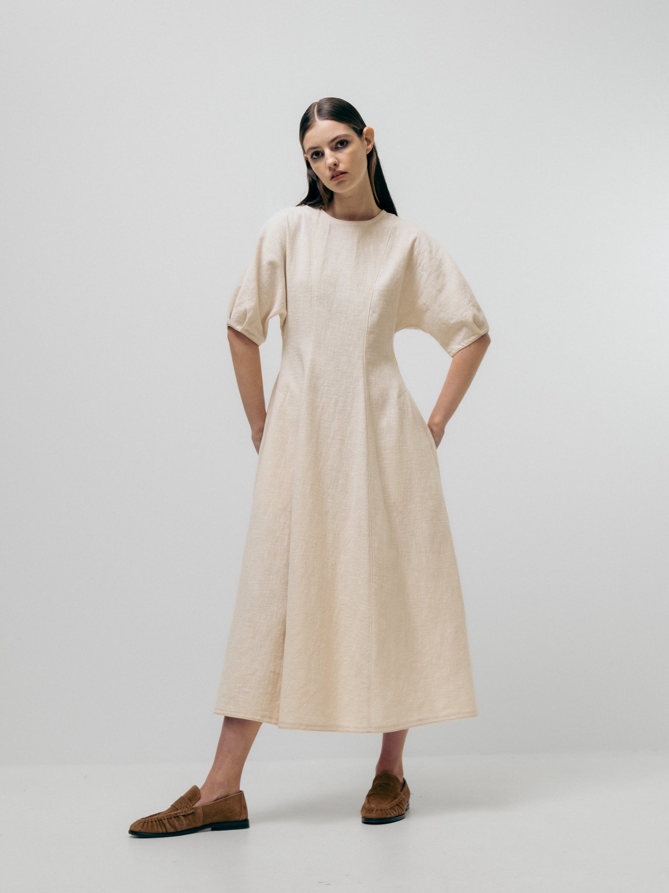 Against a plain backdrop, a person models the Emin and Paul Structured Dress - Cream. This pure linen A-line dress has short sleeves, is light-colored for elegance, and is paired with brown shoes for a simple yet chic look.