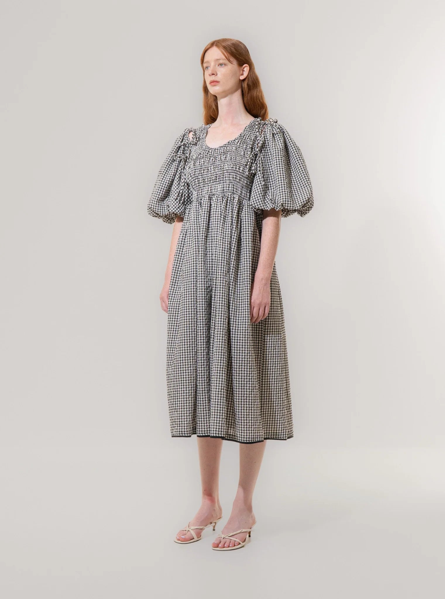 A woman wears the Emin and Paul Puff Sleeve Tie Dress - Black Check, crafted from 100% cotton, paired with sandals against a plain light background.