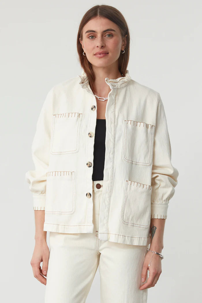 A woman faces forward, wearing the Seventy & Mochi Pablo Denim Jacket in ecru with large front pockets, styled over a black top and matching off-white pants.