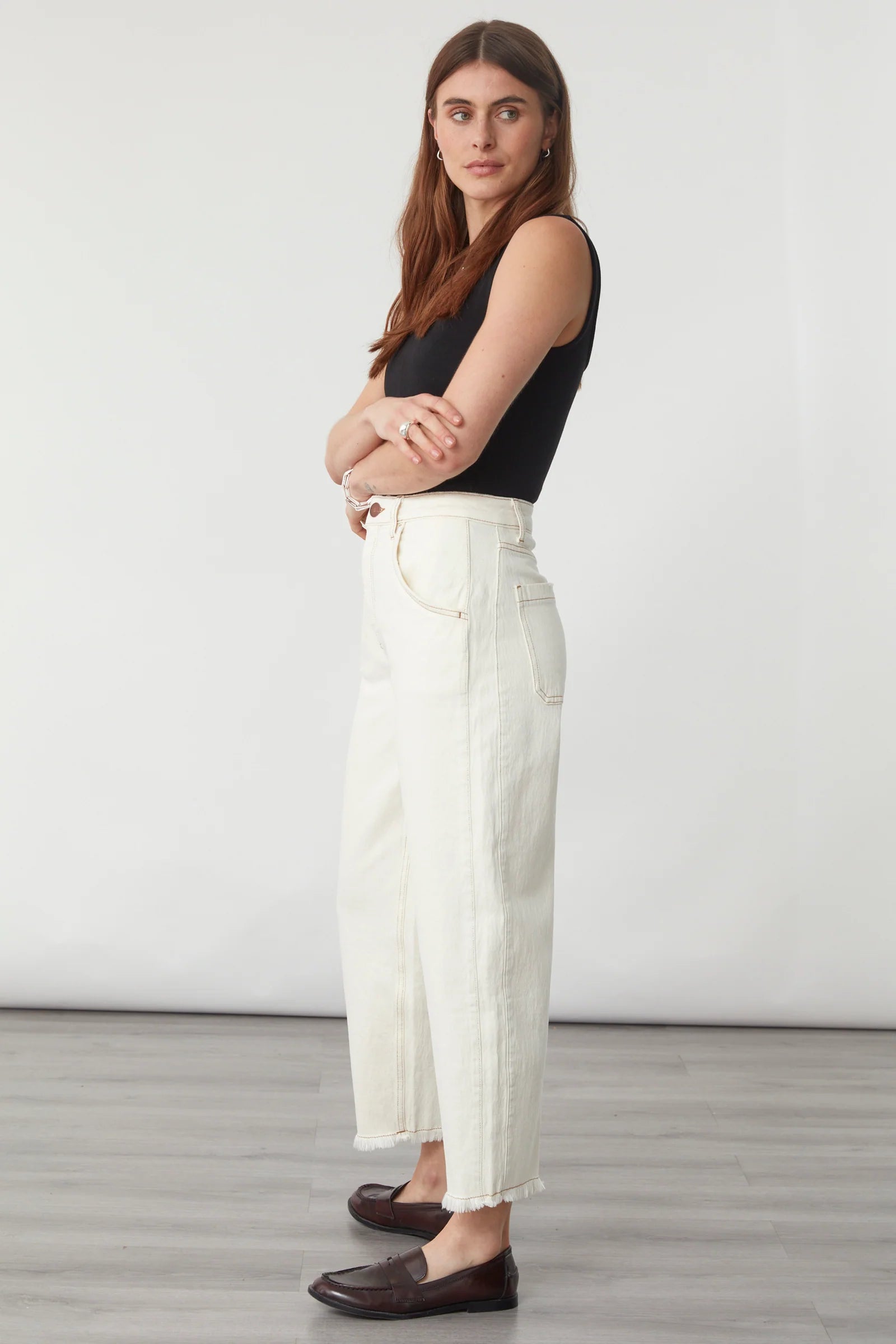 A woman stands sideways against a plain white background on a light wood floor, wearing Seventy & Mochi’s Bo Barrel Leg Jeans in ecru—a super high-rise rigid denim style with a barrel leg silhouette—paired with a black sleeveless top and brown loafers.
