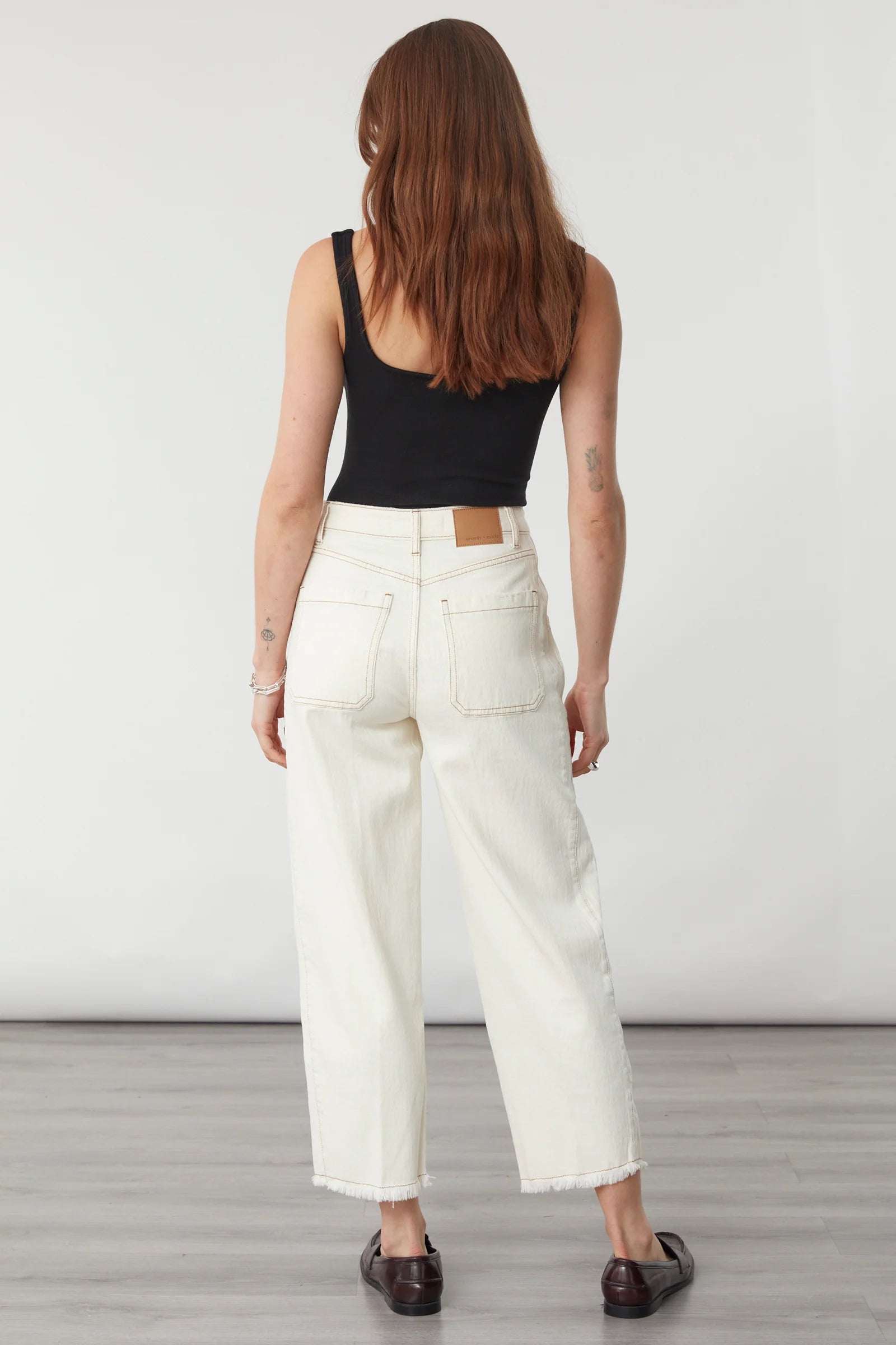 A person with long brown hair wearing a black tank top, dark shoes, and Seventy & Mochi’s Bo Barrel Leg Jeans in Ecru—featuring a super high-rise waist and rigid denim—stands on a gray wooden floor facing away from the camera.