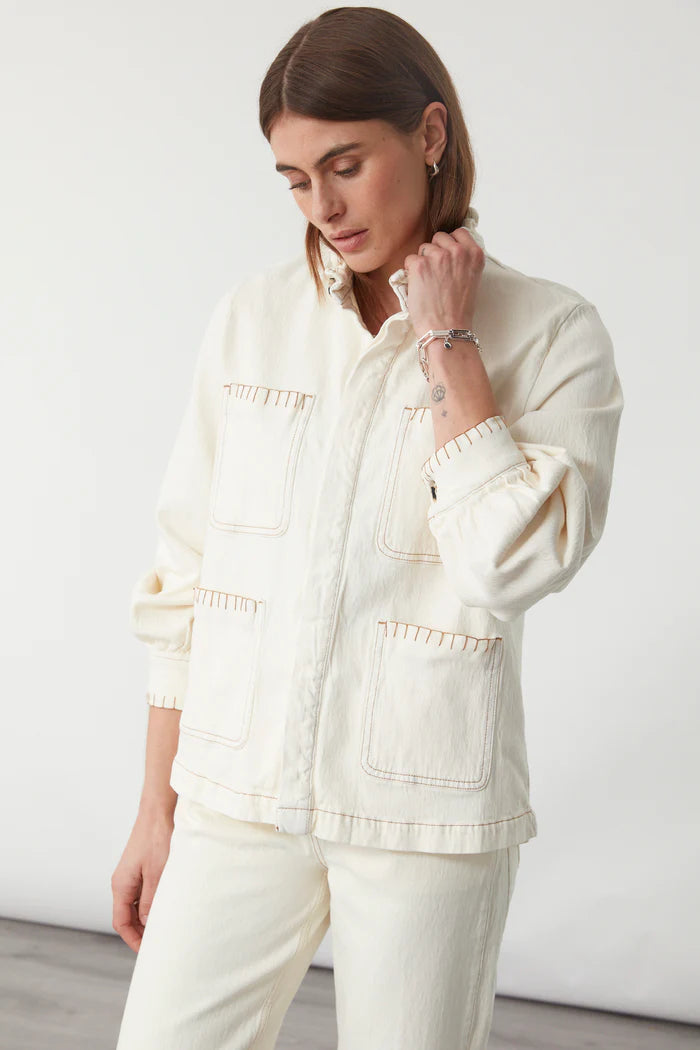 A woman in the Seventy & Mochi Pablo Denim Jacket - Ecru, featuring large front pockets, stands against a plain background in matching pants, gazing downward with one hand touching her frill collar.